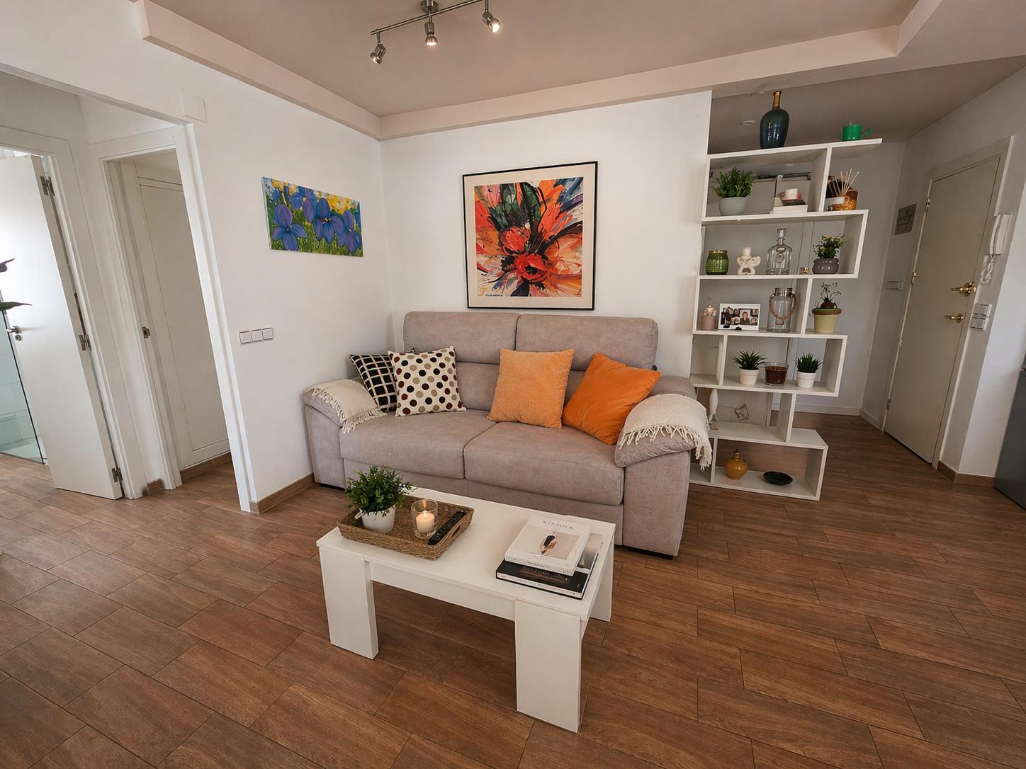 Gezellige zithoek met stoffen bank, witte salontafel en keramische vloer met houtlook. Een geometrische open kast scheidt de woonkamer van de hal.