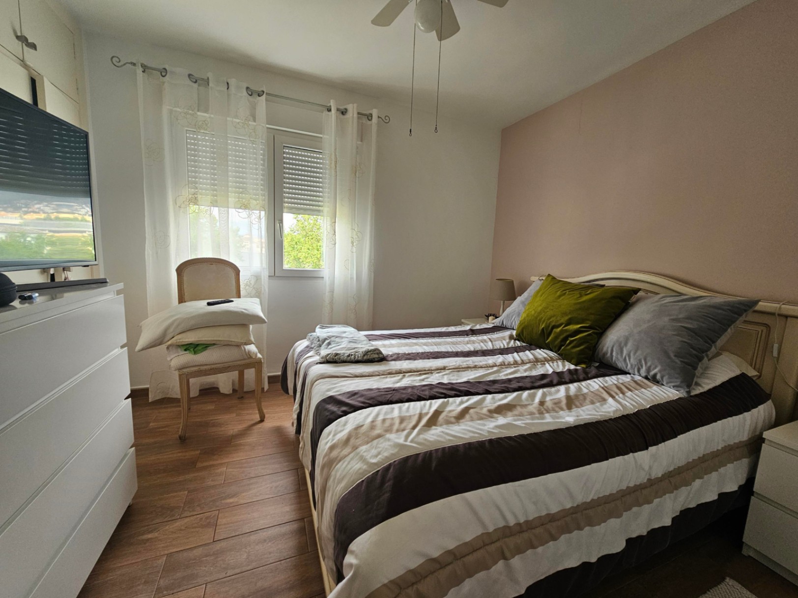 Lichte slaapkamer met een roze accentmuur, vloertegels met houtlook, een plafondventilator en een raam voorzien van rolluiken en vitrage.