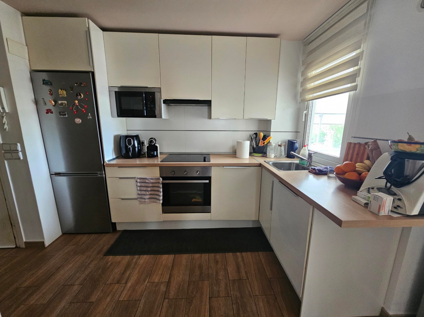 Moderne L-vormige keuken met witte kasten, licht houten werkblad en roestvrijstalen apparatuur, voorzien van een keramische vloer met houtlook.
