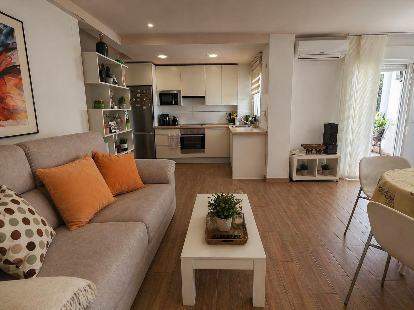 Open woonkamer met houtlook tegelvloer, moderne witte keuken voorzien van inbouwapparatuur en directe toegang tot het balkon via een glazen schuifdeur.
