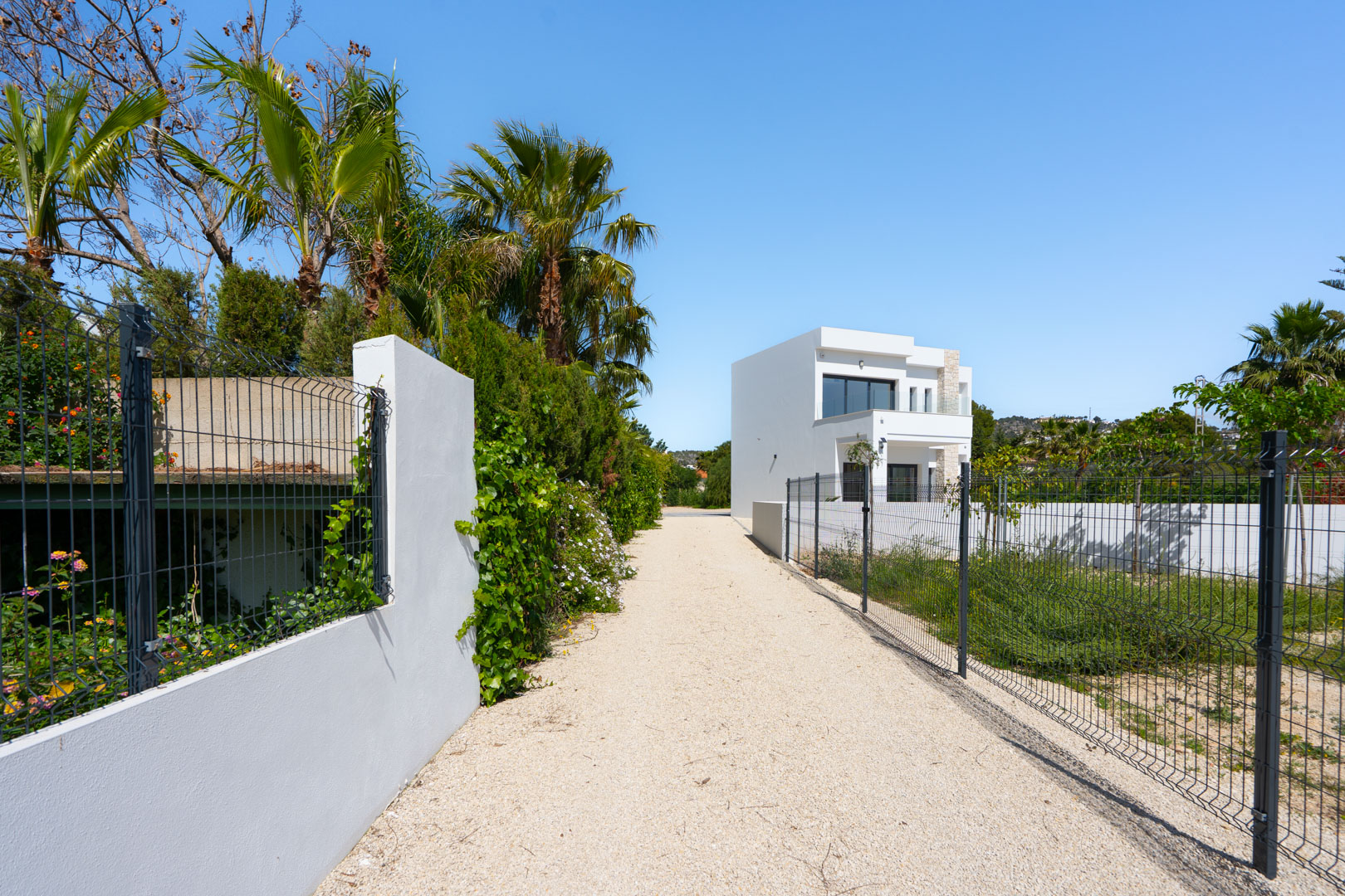 Lange oprit van grind naar de moderne witte villa, omzoomd door hekwerk, mediterrane palmbomen en een uitgestrekt vlak perceel.