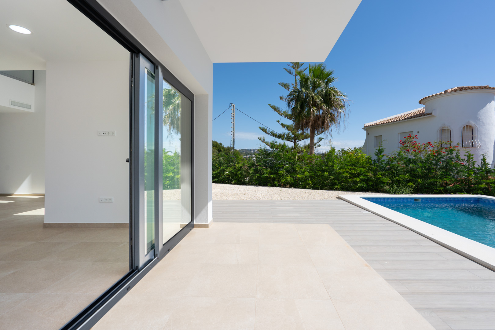 Overdekt terras met porseleinen vloertegels en grote schuifdeuren die toegang bieden tot het zwembad met houtlook terrasplanken en mediterrane beplanting.