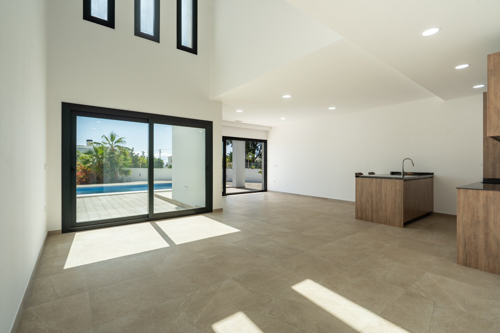 Woonkamer met dubbele hoogte, open designkeuken met kookeiland, porseleinen vloeren en grote schuiframen die toegang bieden tot het zwembad.