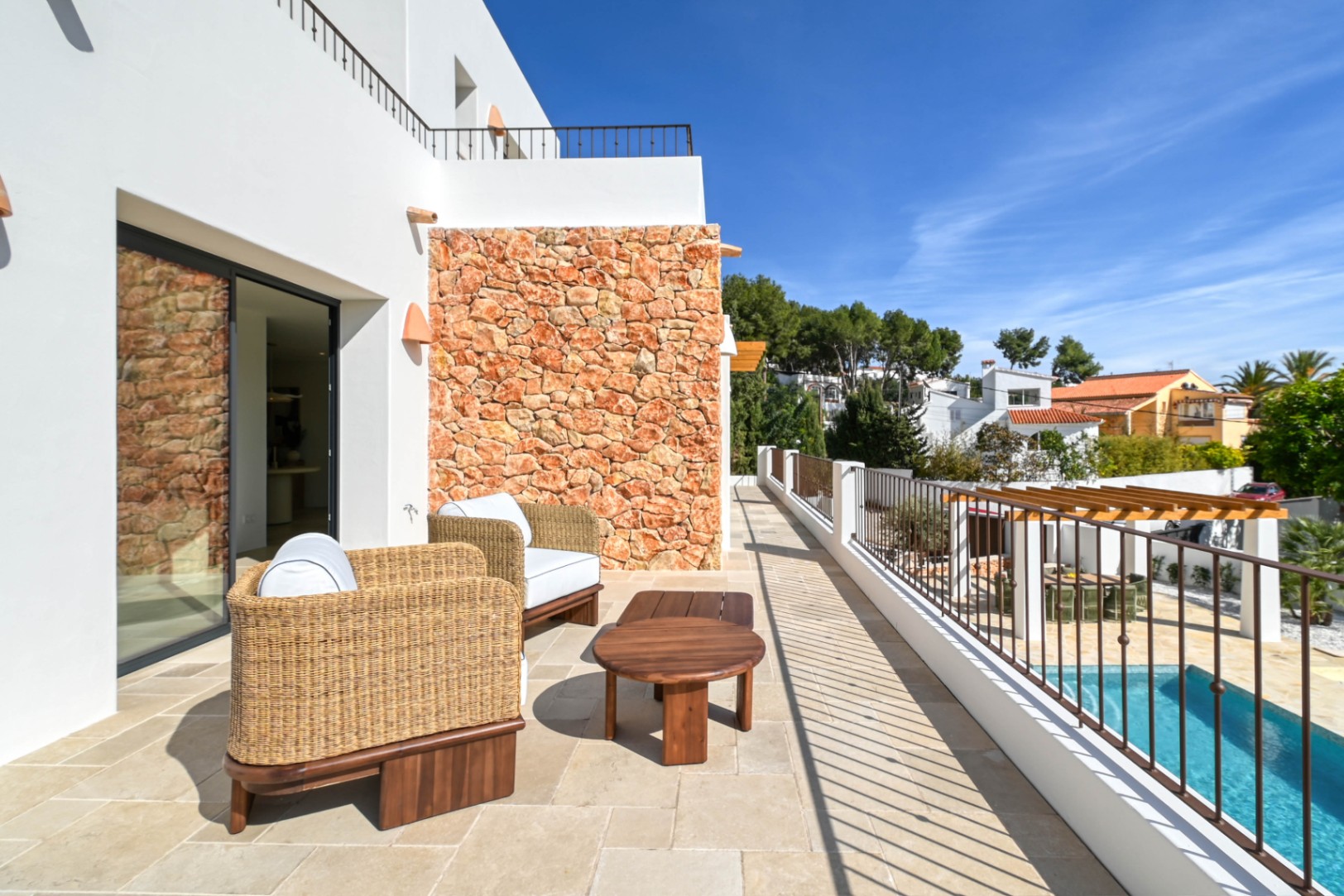 Bovenste terras met natuurstenen tegels, rieten fauteuils en een wand van natuursteen, met uitzicht op het zwembad en de lager gelegen eethoek onder de pergola.