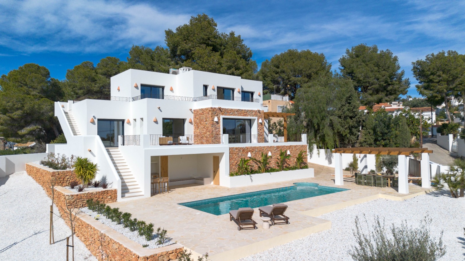 Buitenaanzicht van de moderne villa met witte gevels, natuurstenen muren, een rechthoekig zwembad en meerdere terrassen omgeven door mediterrane beplanting.