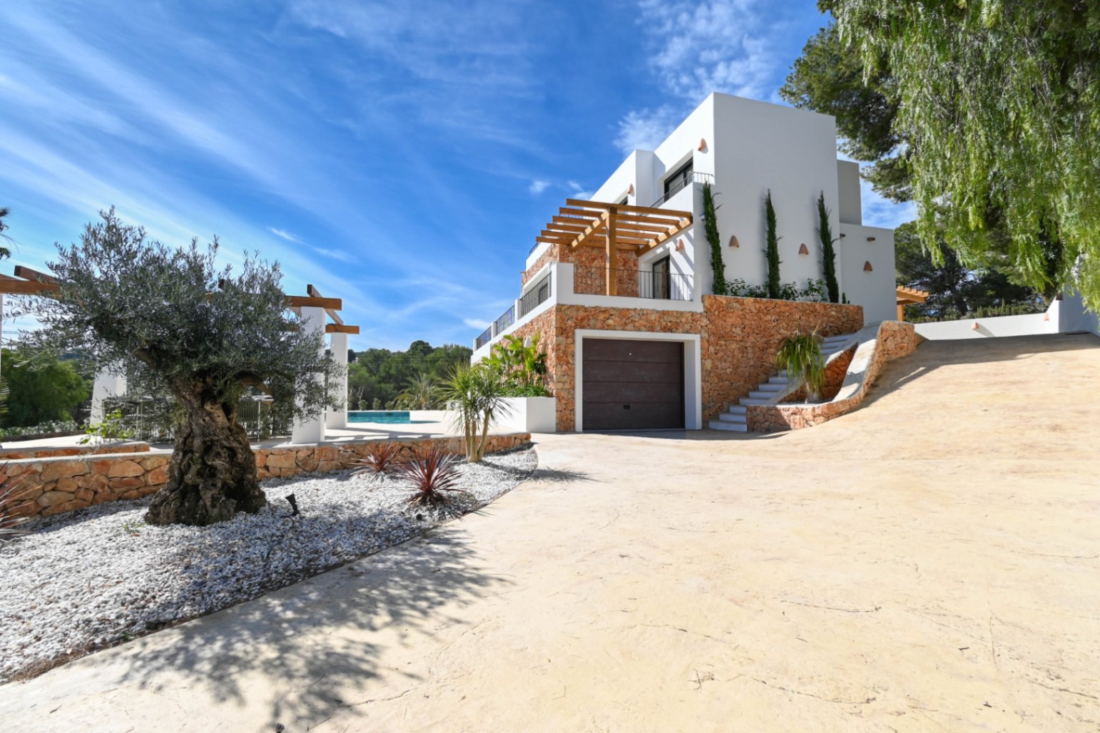 Buitenaanzicht van de villa met natuurstenen gevel, geïntegreerde garage en een oprit omgeven door een olijfboom en mediterrane beplanting.