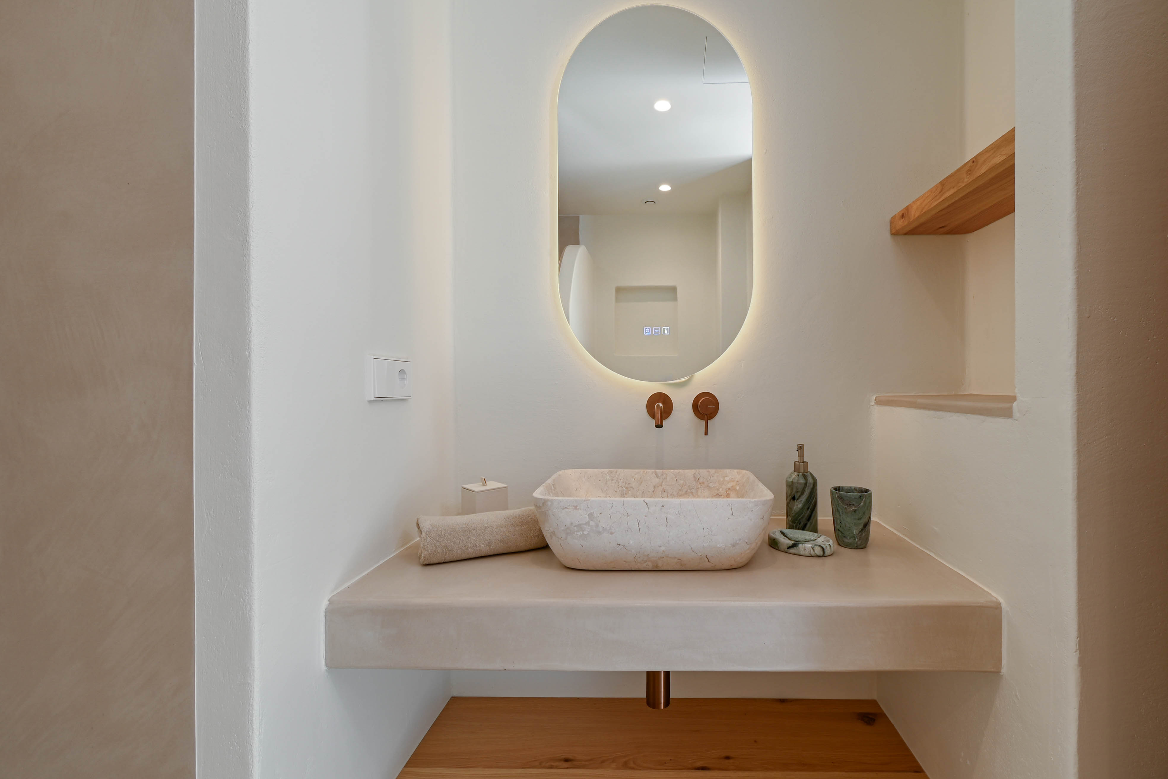 Designbadkamer met natuurstenen waskom op een blad van microcement, koperen inbouwkranen en een ovale verlichte spiegel met houten accenten.