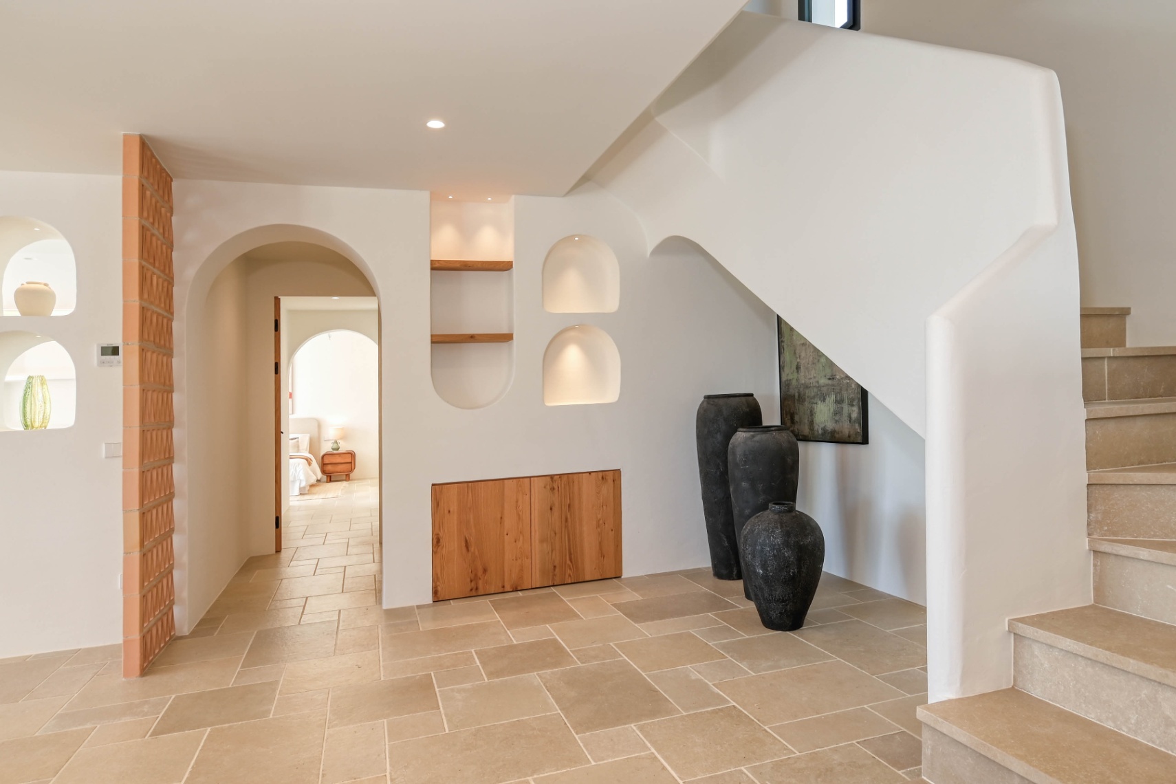 Hal in mediterrane stijl met stenen trap, verlichte nissen en geïntegreerde houten kasten. De natuurstenen vloer loopt door onder elegante bogen naar de kamers.