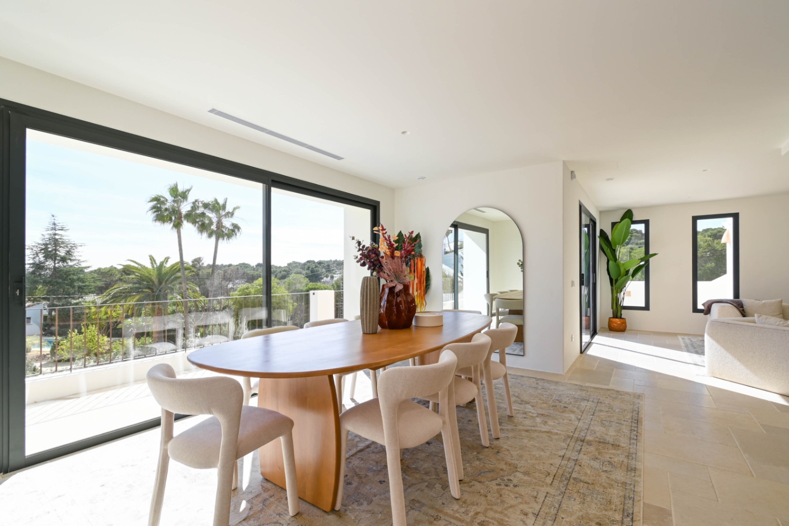 Lichtrijke eethoek met een ovale houten tafel en grote glazen schuifdeuren die uitkijken op palmbomen en de omliggende mediterrane natuur.