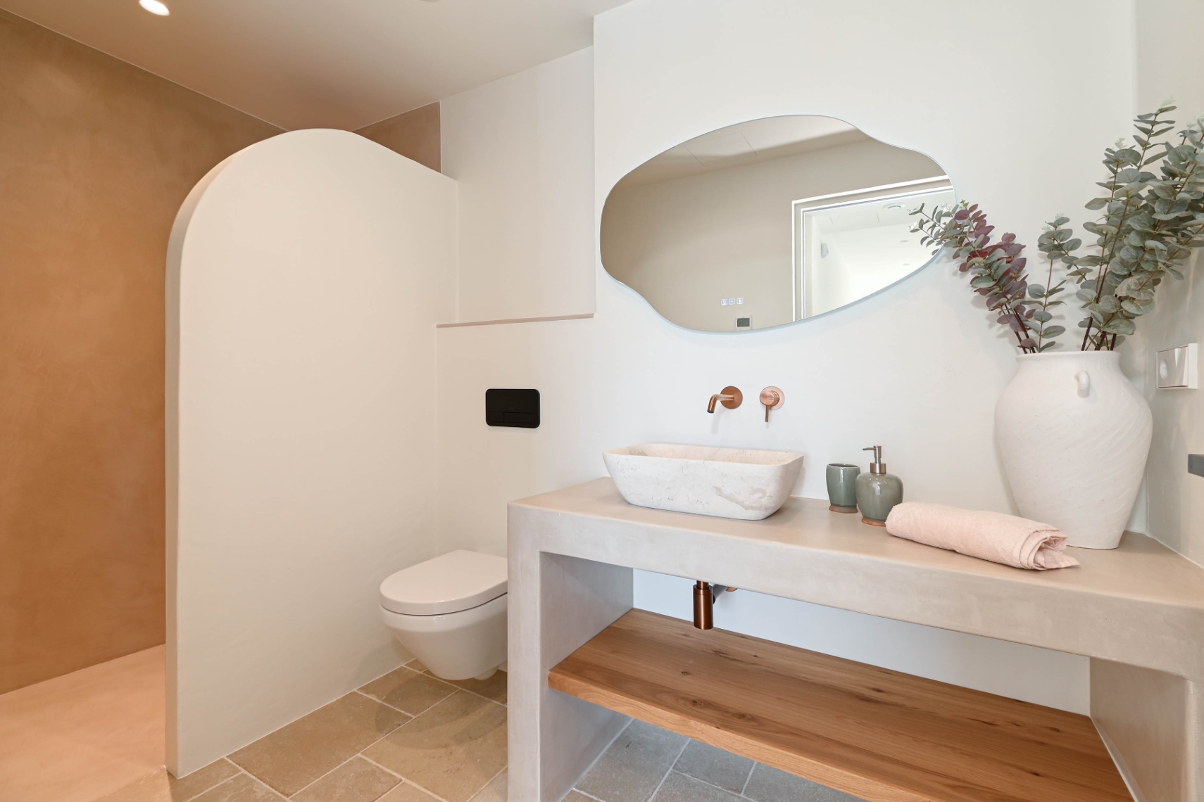 Moderne badkamer met een stenen waskom op een microcement meubel, koperen inbouwkranen, een organisch gevormde spiegel en een stijlvolle gebogen douchewand.