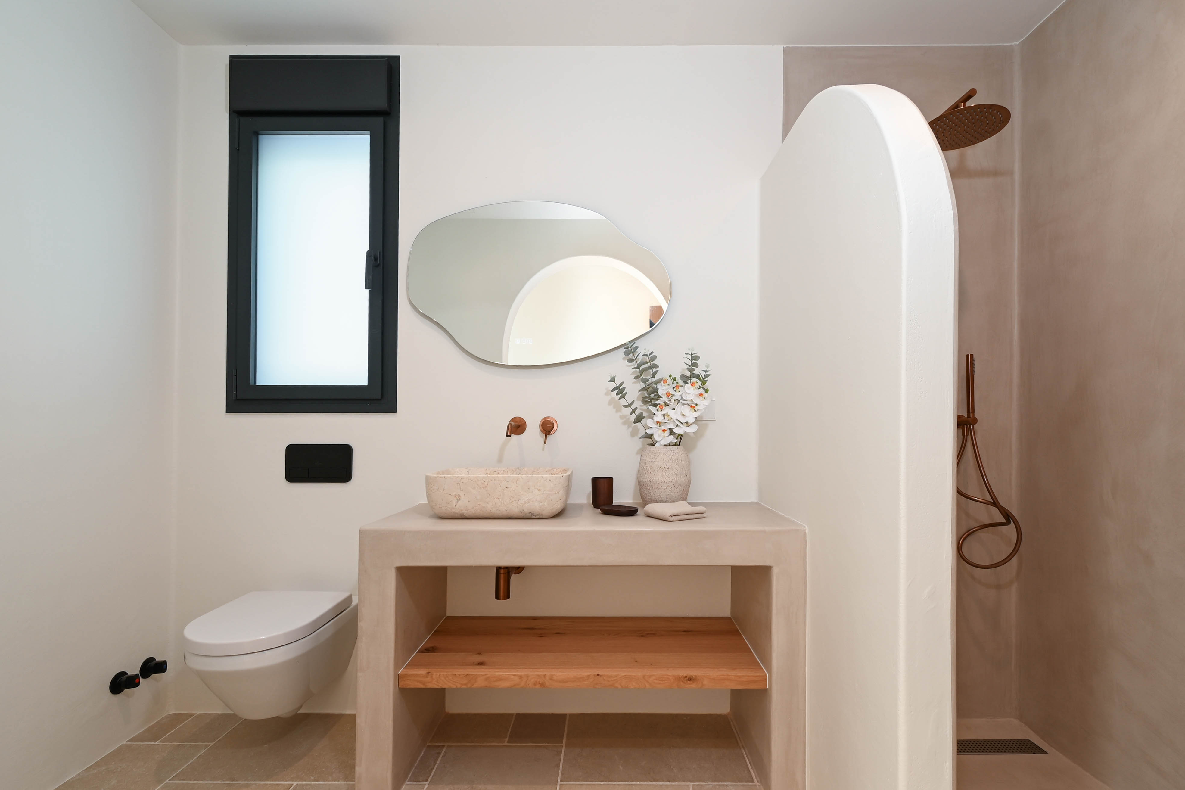 Moderne badkamer met een wastafel van microcement, natuurstenen kom en koperen kranen. Voorzien van een organische spiegel, hangtoilet en inloopdouche met een gebogen tussenwand.