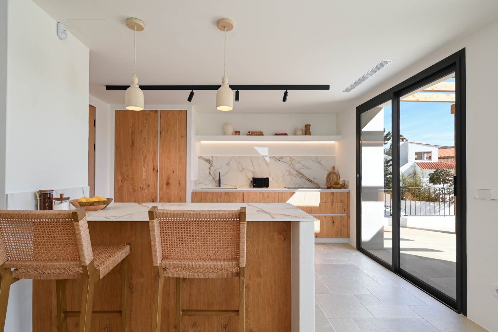 Open keuken met houten eiland, marmerlook aanrechtbladen en rieten krukken, verbonden met het terras via grote schuifdeuren met zwart profiel.