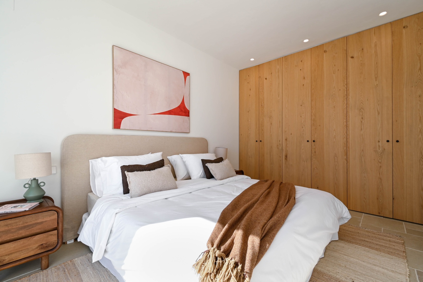 Slaapkamer met kamerhoge inbouwkasten van natuurlijk hout, een beige gestoffeerd hoofdbord en inbouwspots. De ruimte is afgewerkt met een tegelvloer en een houten nachtkastje.
