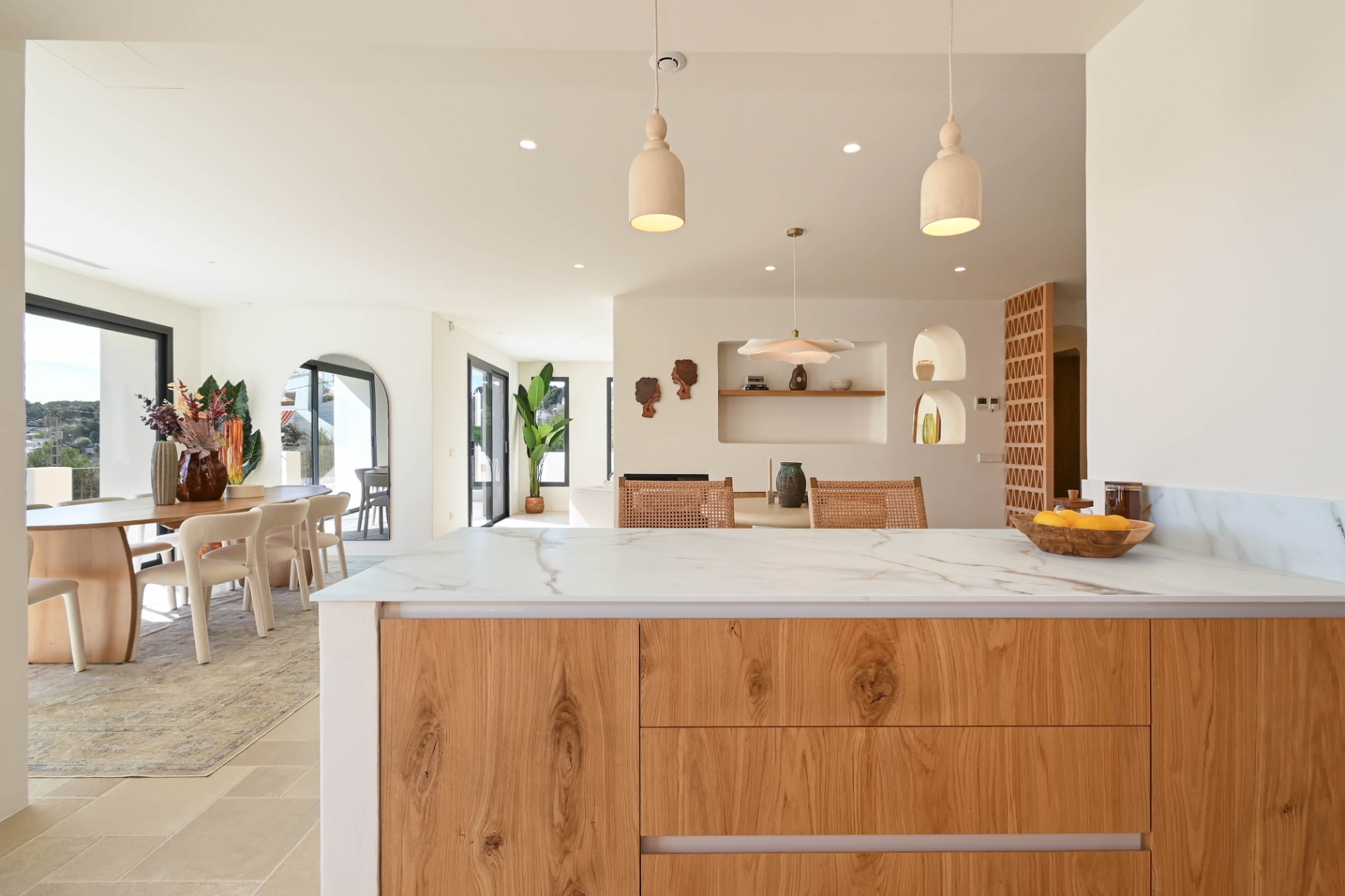 Zicht vanaf het kookeiland met natuurstenen blad op de open eetkamer, voorzien van decoratieve nissen, houten afwerkingen en grote glazen schuifpuien.