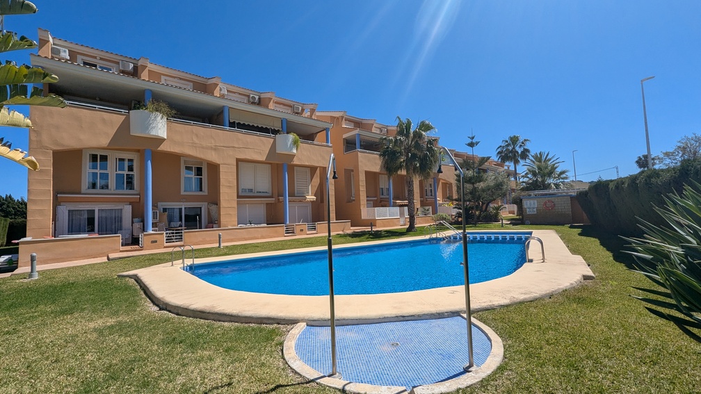 Gemeenschappelijk niervormig zwembad omgeven door een verzorgd gazon en mediterrane tuinen, met het terracotta-kleurige appartementengebouw en blauwe zuilen op de achtergrond.