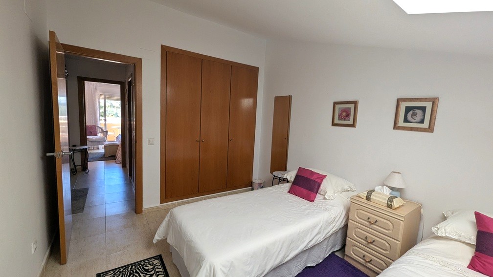 Slaapkamer met twee eenpersoonsbedden, houten inbouwkasten en een tegelvloer. De open deur biedt toegang tot de gang en een lichte kamer met terras.