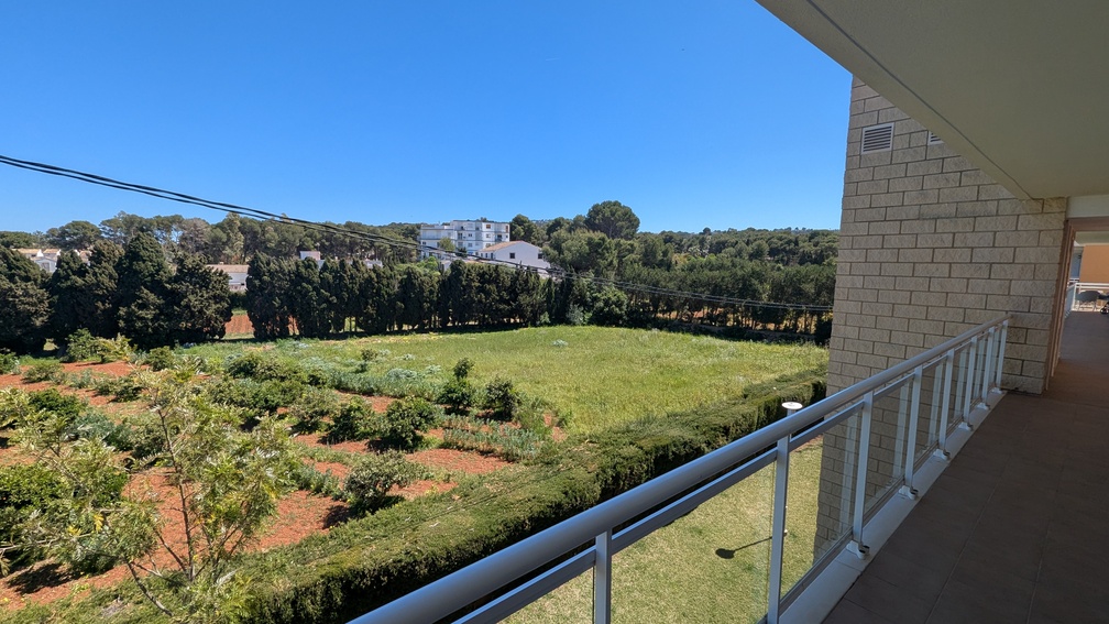 Uitzicht vanaf het betegelde terras met glazen balustrade op open groene landschappen en een rij cipressen onder een heldere hemel.