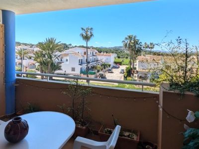 Balkon met tafel en stoel, met vrij uitzicht op een woonwijk met witte huizen, palmbomen en groene heuvels onder een helderblauwe lucht.