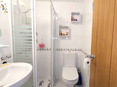 De moderne doucheruimte heeft wit betegelde muren met een decoratieve rand, een glazen douchecabine met matte strepen, een wandwastafel en een toilet. Twee vierkante wandplanken zijn aanwezig.