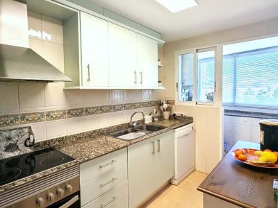 Volledig uitgeruste keuken met lichte kasten, graniet-effect werkbladen, geïntegreerde kookplaat en oven, en een raam naar een aangrenzende bijkeuken.