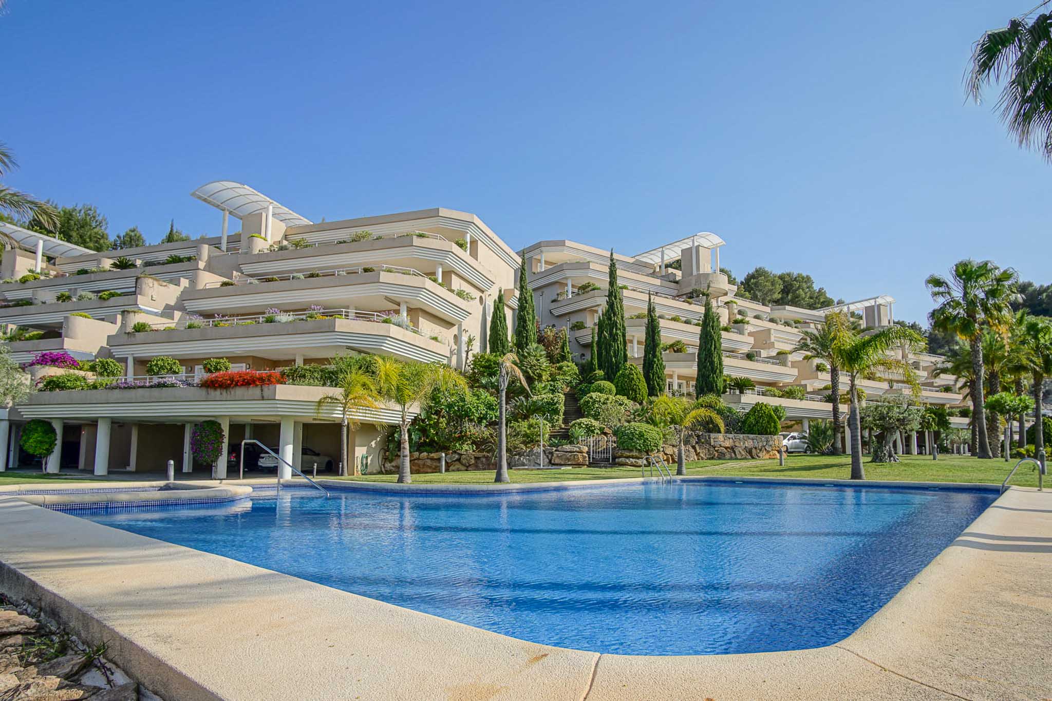 Exterieur van het Las Mimosas-complex met een groot gemeenschappelijk zwembad en terrasvormige architectuur te midden van mediterrane tuinen en palmbomen.