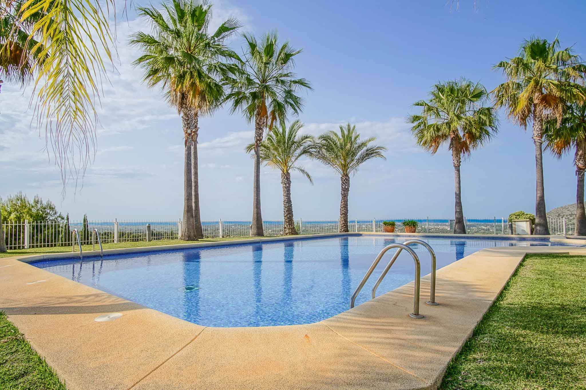 Groot gemeenschappelijk zwembad met blauwe tegels, omgeven door een stenen zonneterras, gazon en palmbomen met panoramisch uitzicht op de Middellandse Zee.