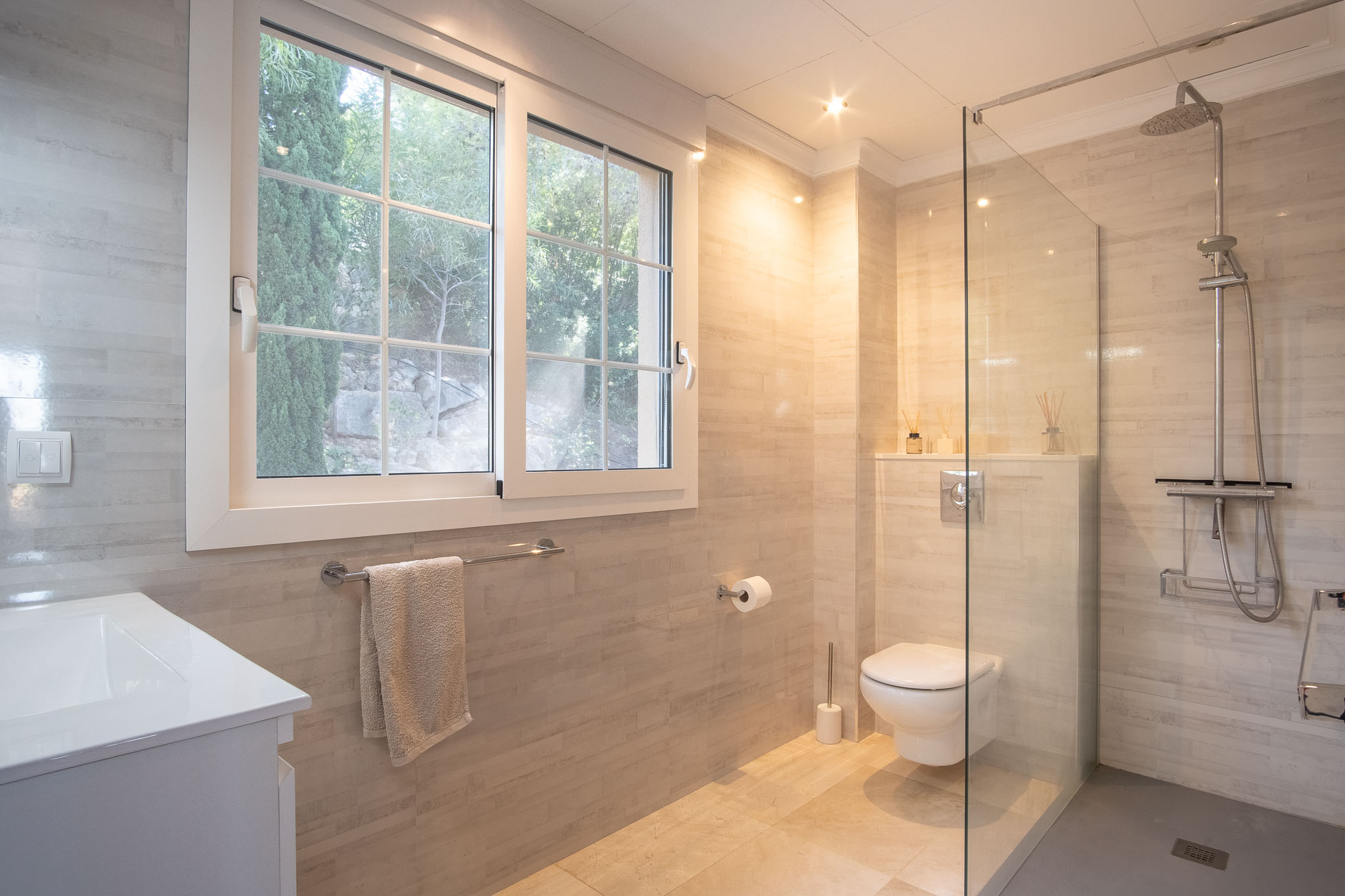 Moderne badkamer met inloopdouche, glazen wand en neutrale betegeling. Voorzien van een hangend toilet, inbouwspots en een groot raam voor natuurlijk daglicht en uitzicht op de tuin.