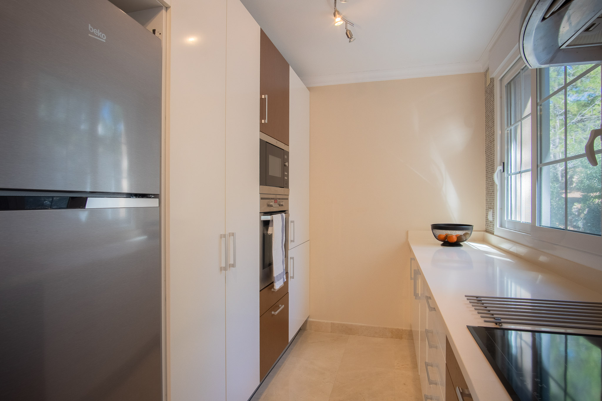Moderne keuken met witte en houten kasten, geïntegreerde roestvrijstalen apparatuur en een wit werkblad. Een zijraam zorgt voor natuurlijk licht bij de inductiekookplaat.