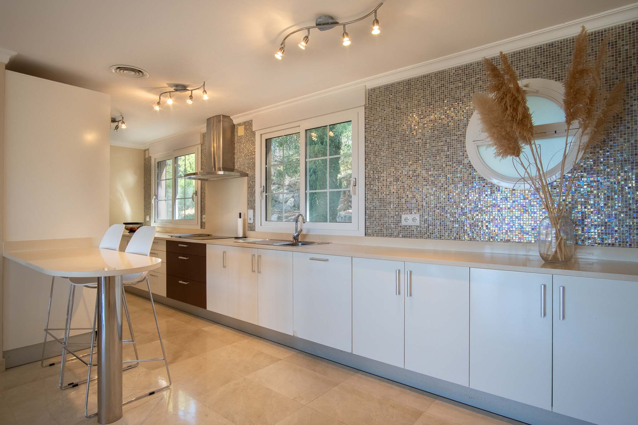Moderne keuken met witte kasten, licht werkblad en een opvallende iriserende mozaïek achterwand. Voorzien van een ontbijttafel, marmeren vloer en natuurlijke lichtinval.