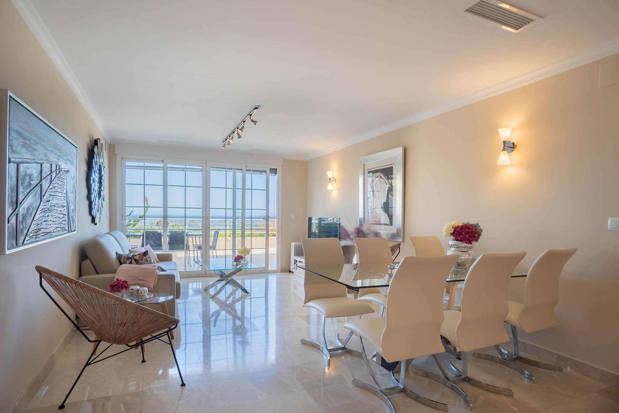 Open woon- en eetkamer met gepolijste marmeren vloeren, modern glazen meubilair en grote schuifdeuren die toegang bieden tot het terras met uitzicht op zee.