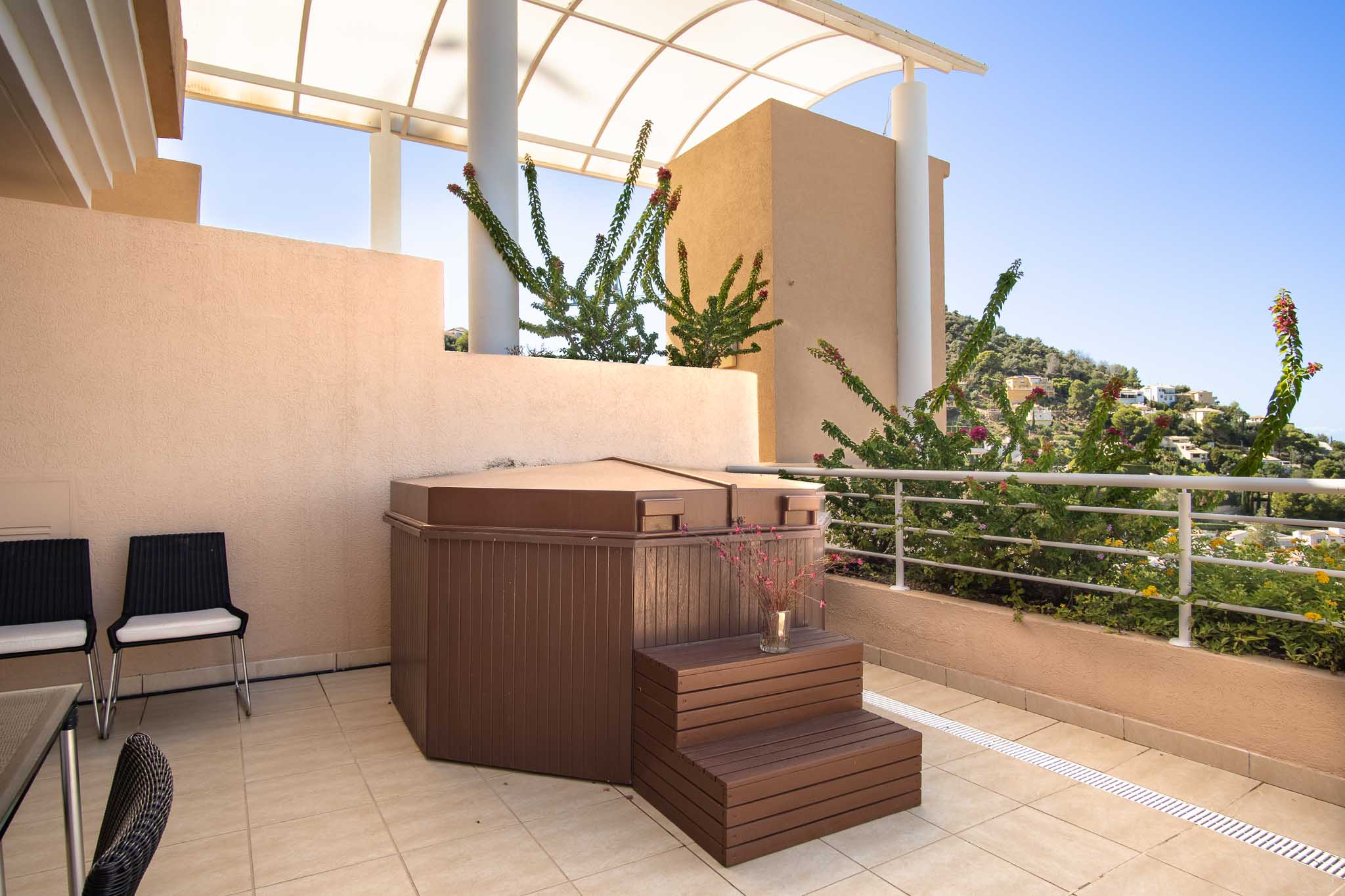 Privéterras met een afgedekte jacuzzi en houten opstapje, gelegen onder een gebogen pergola met uitzicht op de omliggende heuvels en mediterrane beplanting.