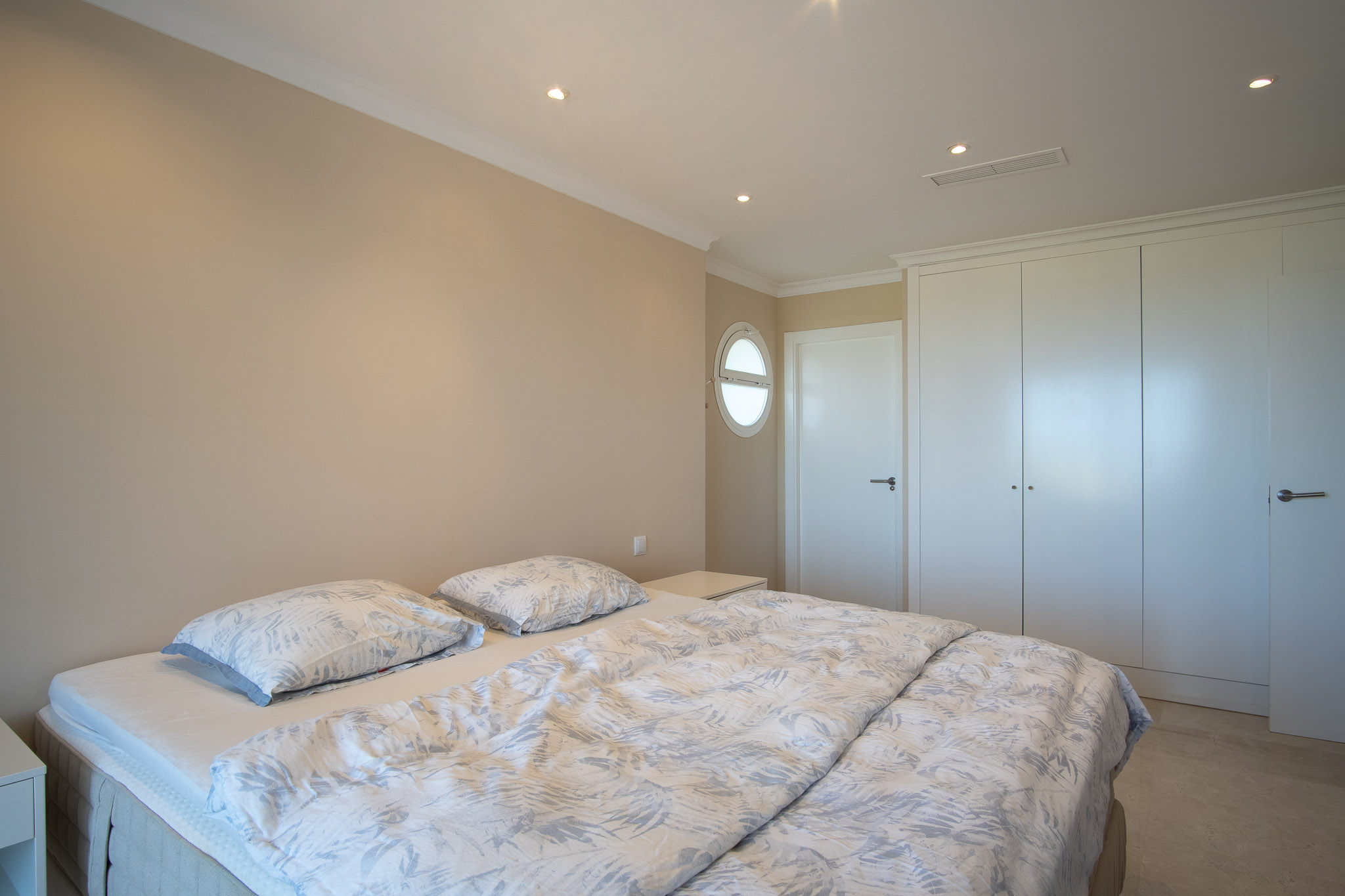 Slaapkamer met tweepersoonsbed, witte inbouwkasten en marmeren vloer. Voorzien van inbouwspots en een karakteristiek rond ossennoogvenster.