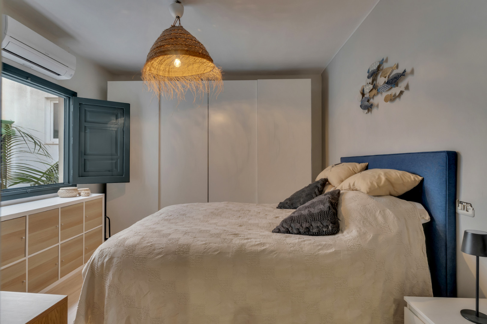 Lichte slaapkamer met blauw hoofdeinde, rieten lamp en een raam met luiken.