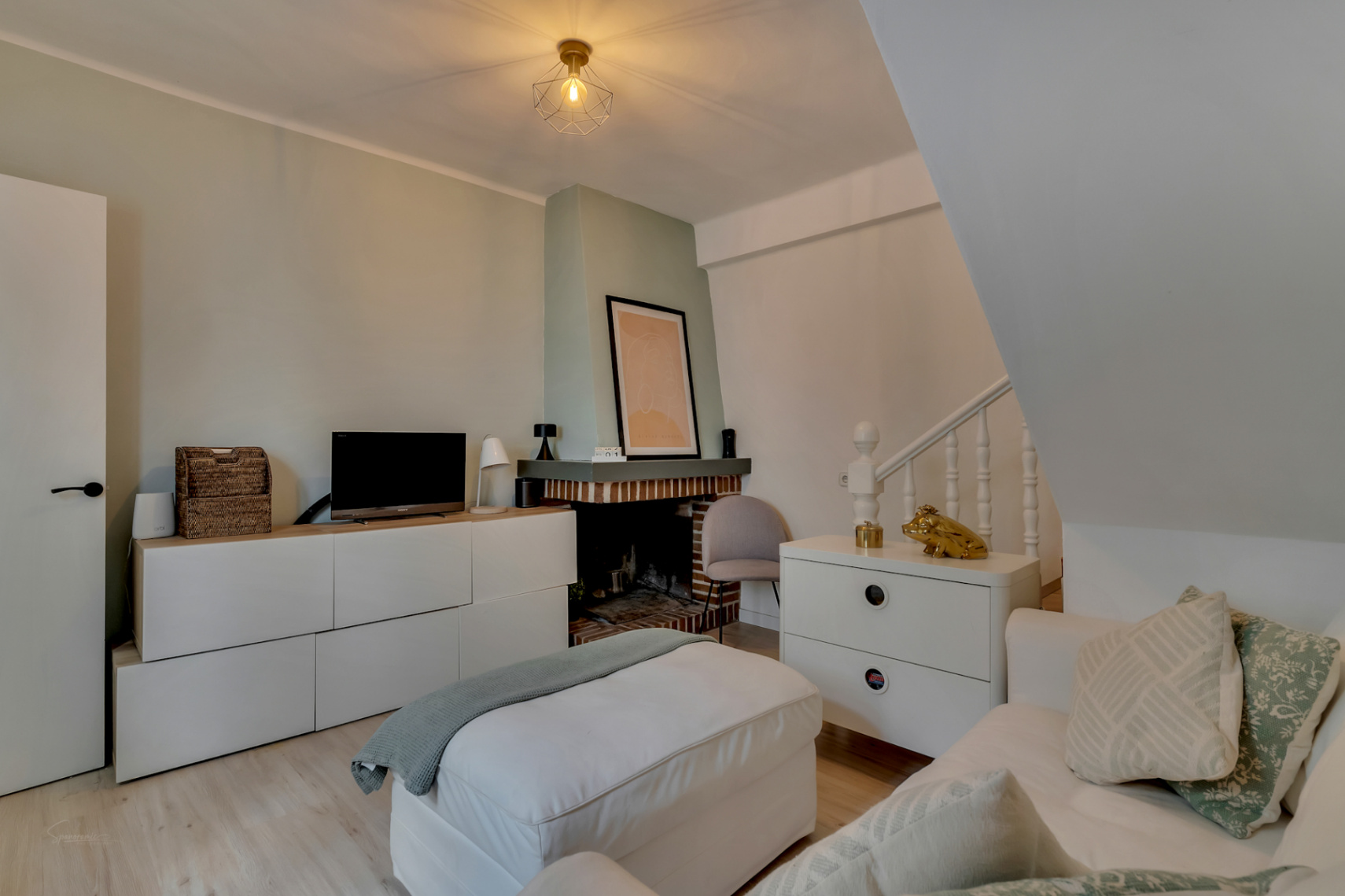Lichte woonkamer met bakstenen open haard, lichte houten vloeren en moderne meubels in neutrale tinten.