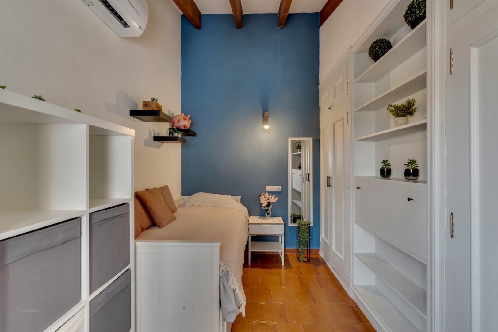 Slaapkamer met blauwe accentmuur, witte stellingkasten en terracotta tegelvloer. Houten balken aan het plafond.