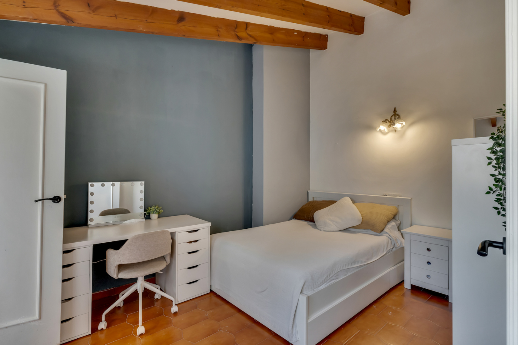 Slaapkamer met terracotta vloer, blauwe accentmuur en wit bureau met verlichte spiegel.
