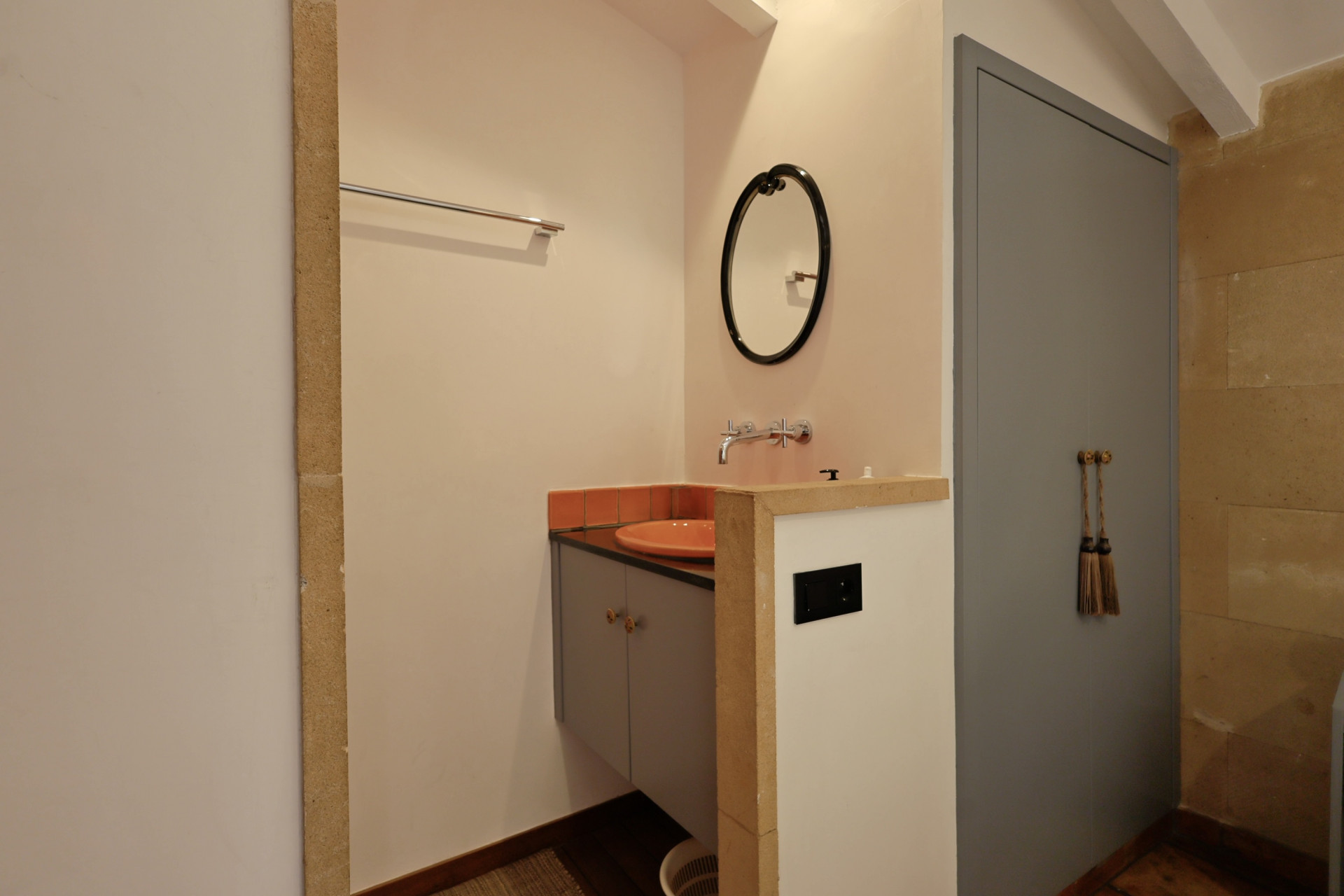 Badkamerdetail met oranje keramische wastafel, ovale spiegel en grijze deur met decoratieve kwasten.
