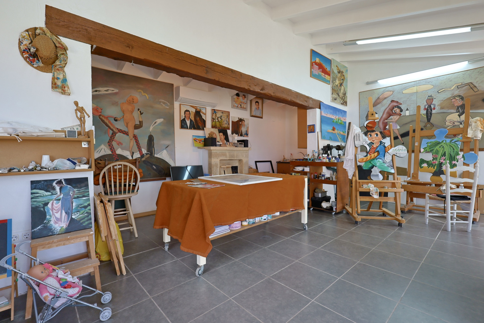 Ruime kunststudio met grijze tegelvloeren, een met oranje stof bedekte tafel en schildersezels met schilderijen.