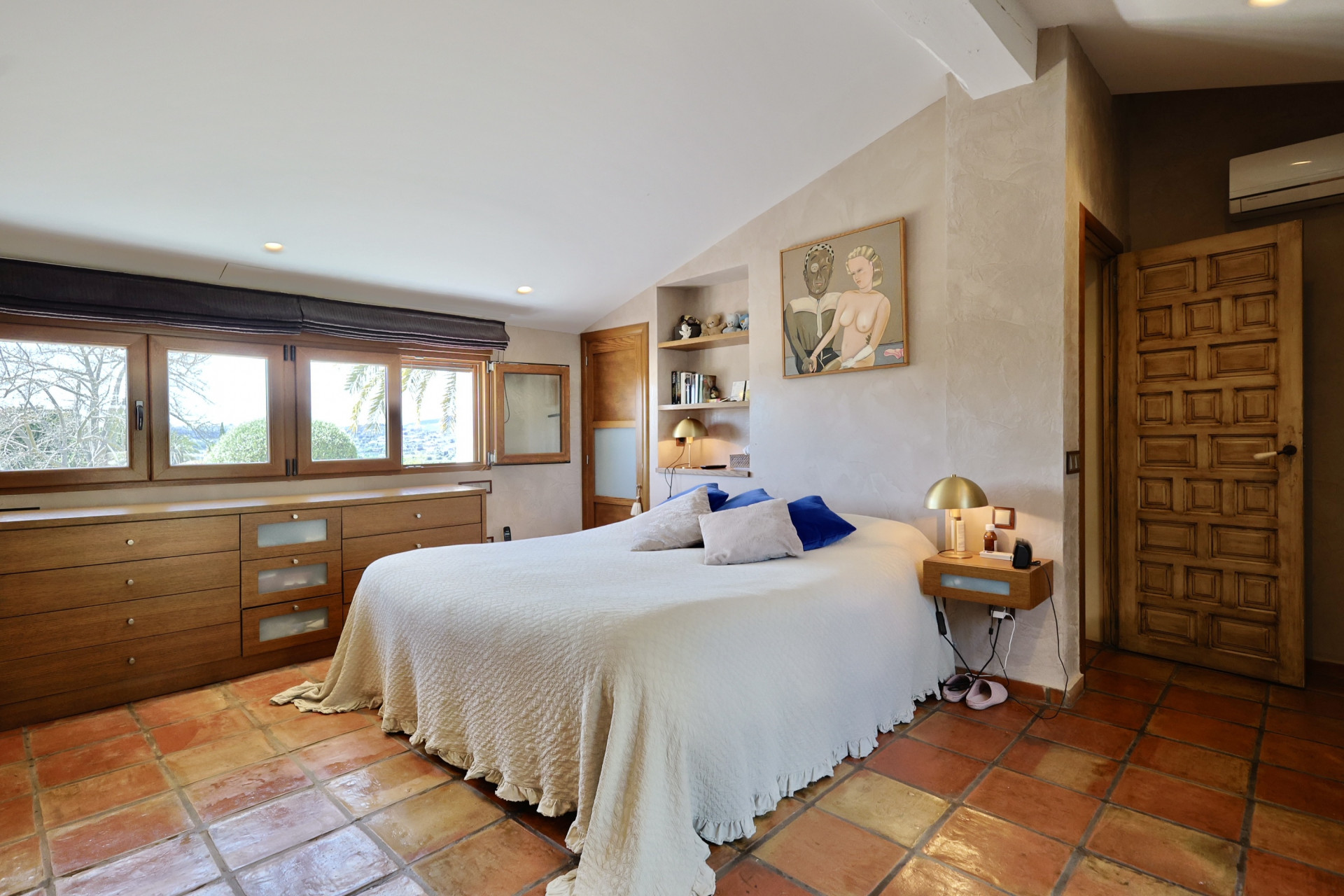 Slaapkamer met terracotta vloertegels, een tweepersoonsbed met wit linnen en een houten hoofdeinde met ramen.