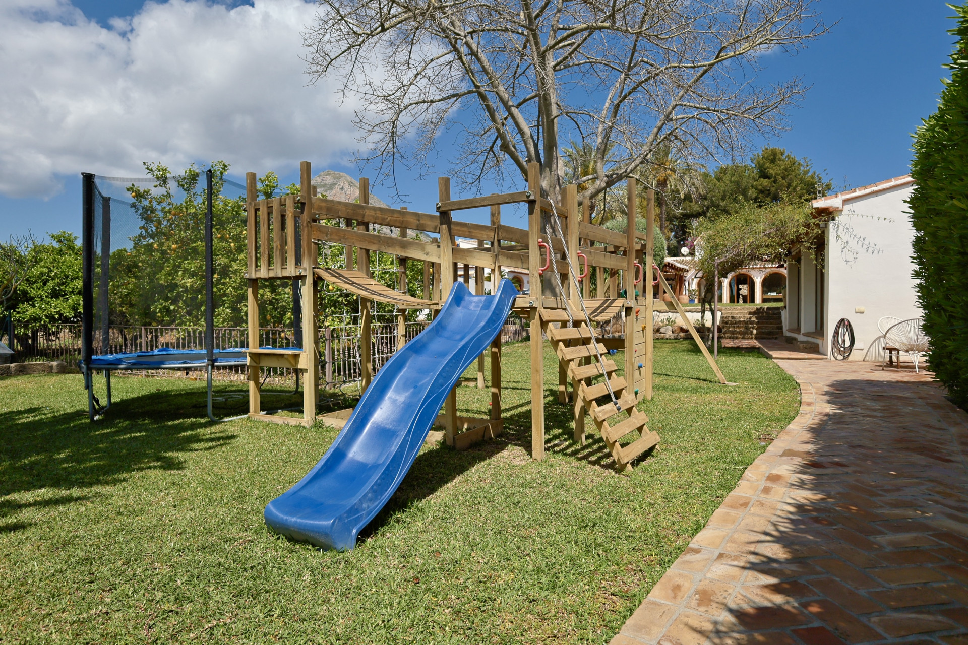Speelplaats met houten structuur, blauwe glijbaan en trampoline in de tuin.