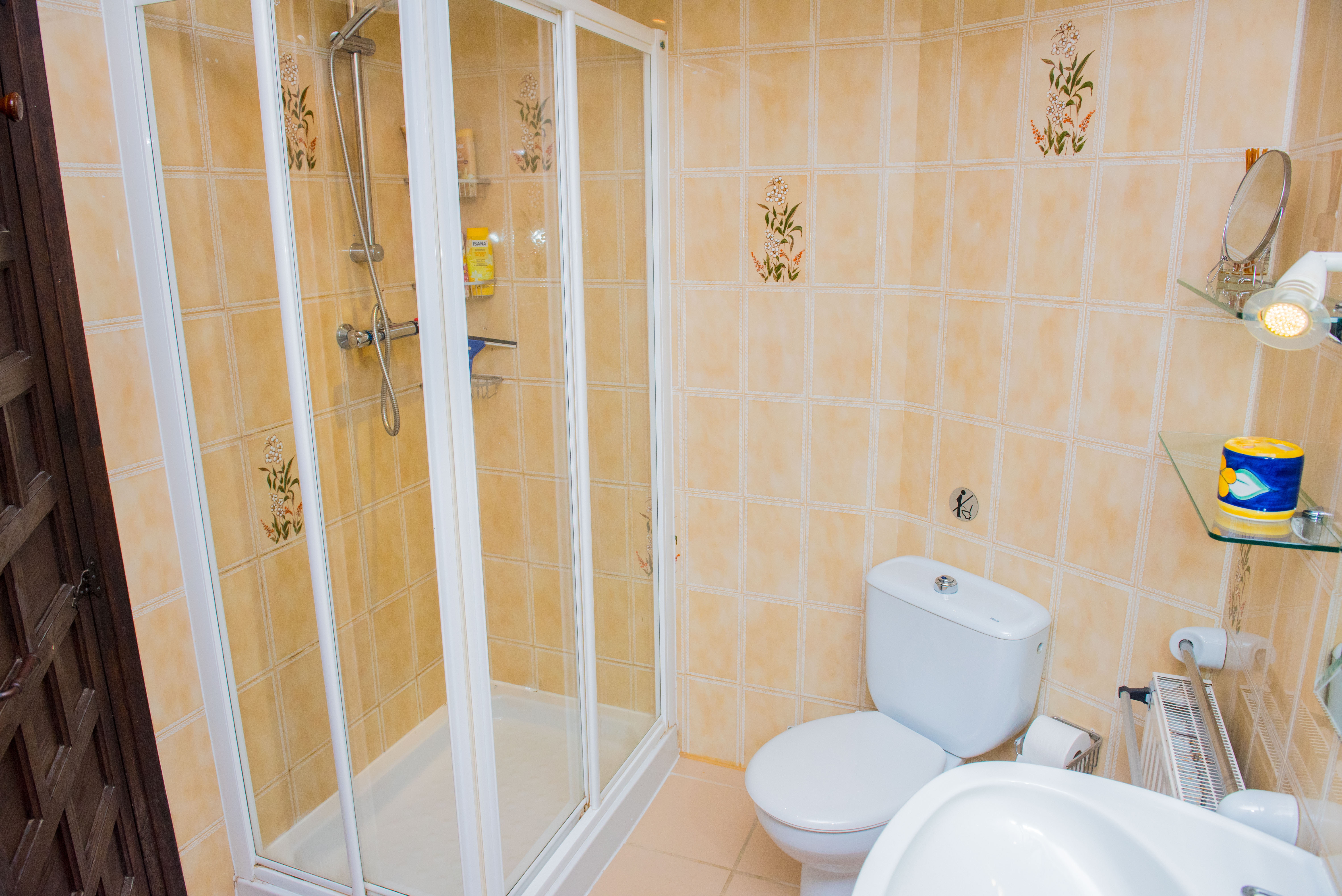 Badkamer met een wit omrande glazen douchecabine, beige wandtegels met florale motieven en wit keramisch sanitair.