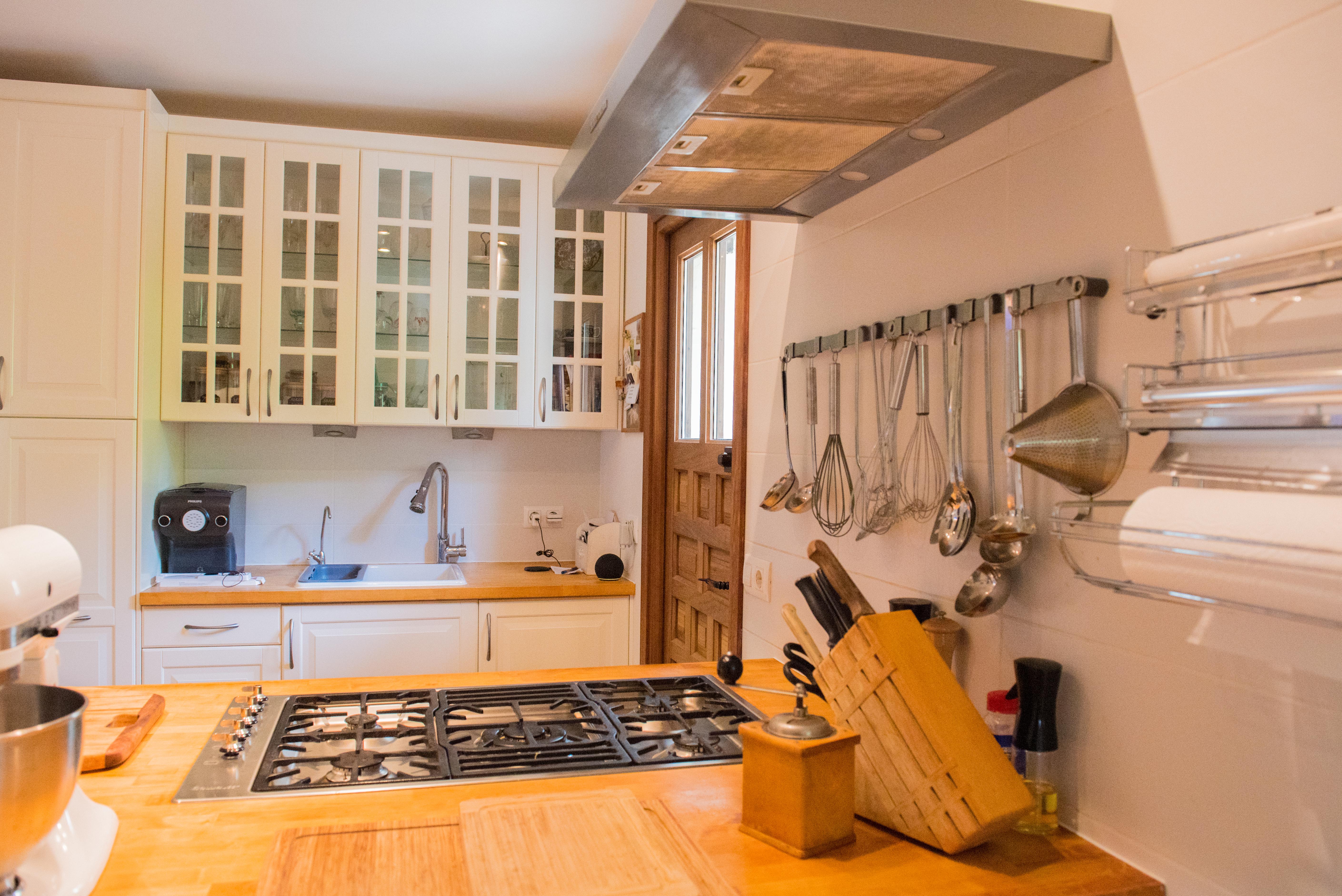 Functionele keuken met houten werkbladen, een geïntegreerde gaskookplaat en witte kasten met glazen bovenfronten. Inclusief werkruimte met hangend keukengerei en een houten deur.