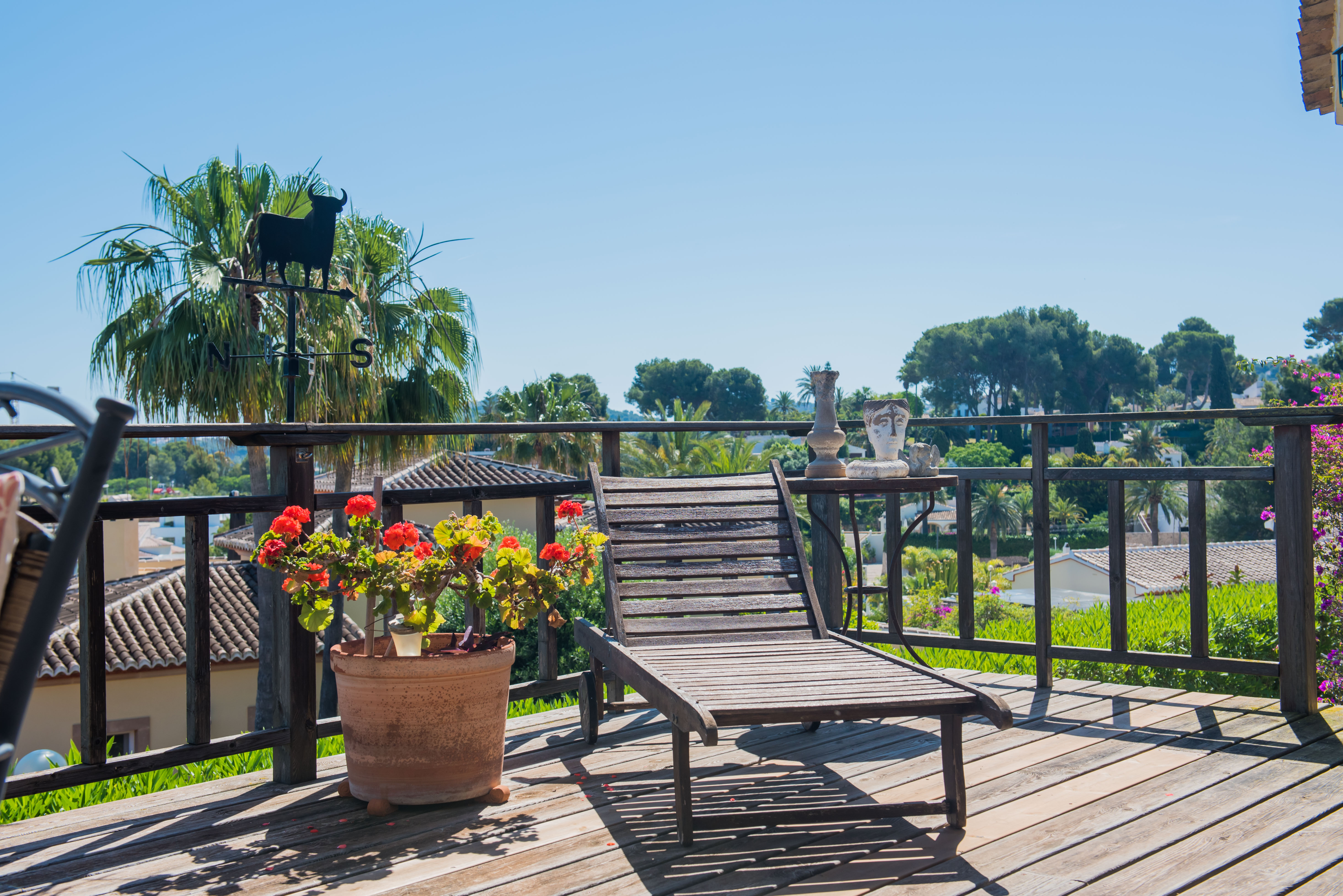 Houten terras met ligstoel, uitzicht op de tuin en decoratieve details zoals een stierenwindvaan en geraniumpotten.