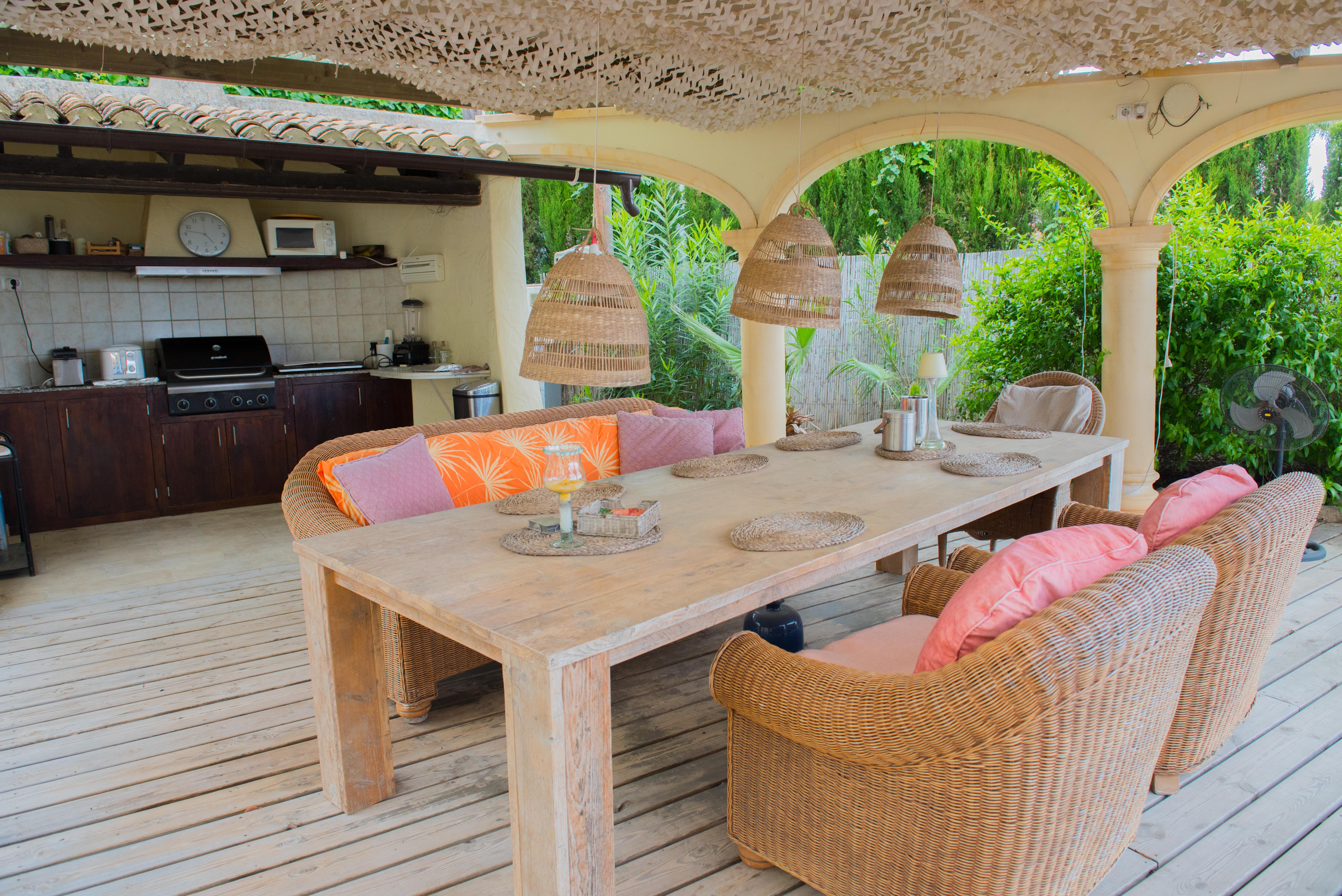 Overdekt terras met een volledig uitgeruste buitenkeuken, massief houten eettafel, rieten fauteuils en decoratieve schaduw op een houten vlonder.