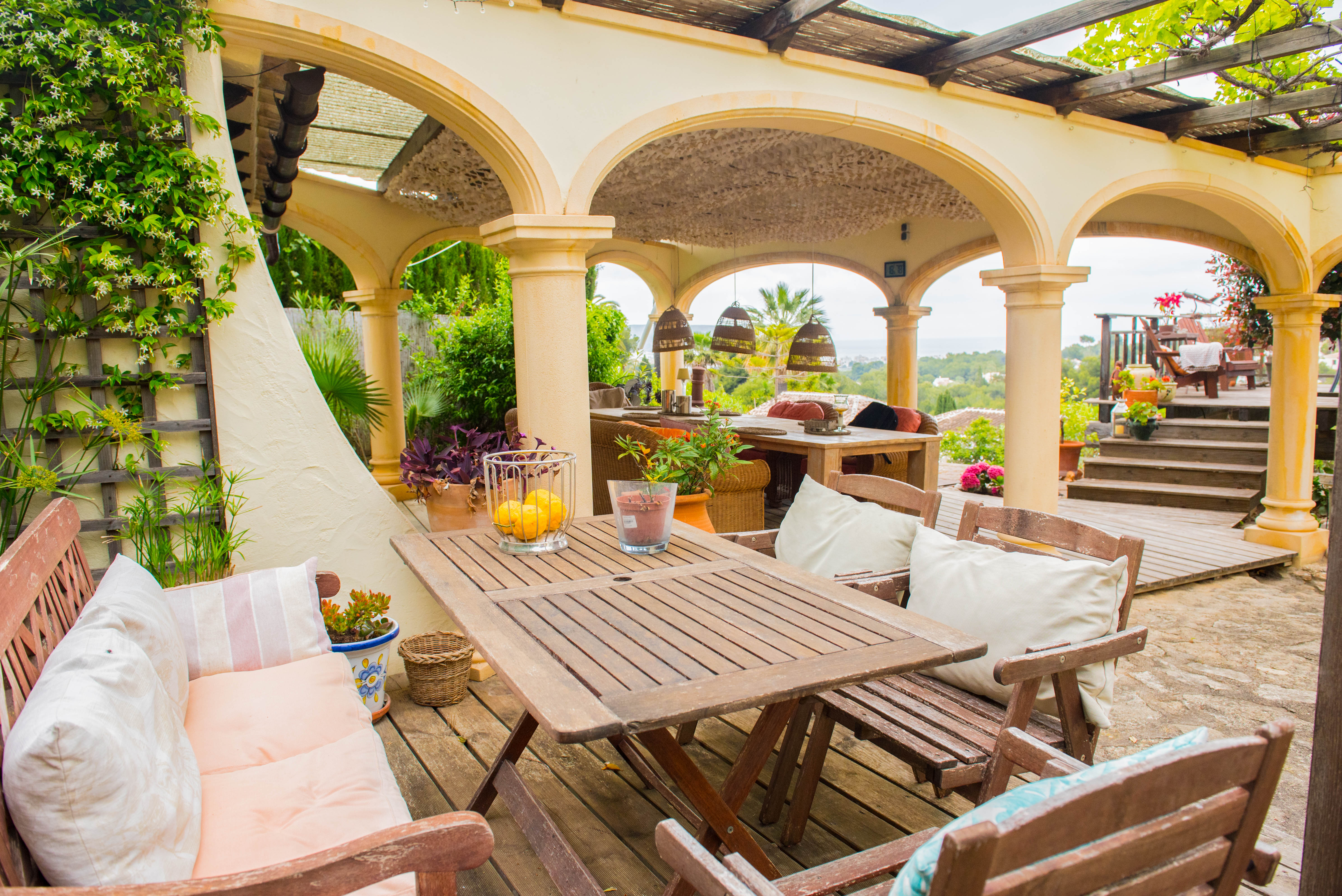 Overdekt terras met mediterrane bogen, houten eethoek, houten vlonders en panoramisch uitzicht op de zee en de groene omgeving.