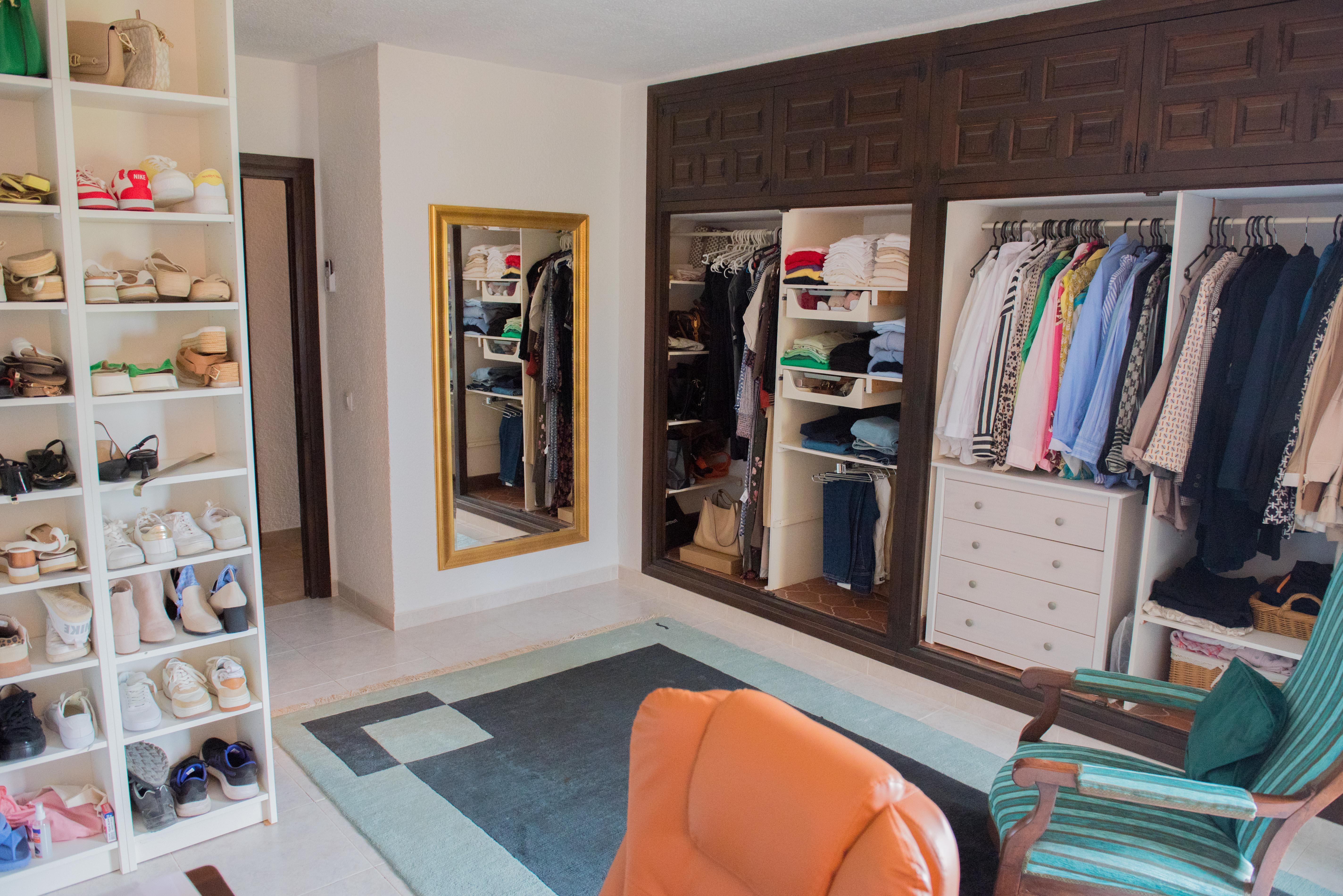 Ruime kleedkamer met ingebouwde kasten van donker hout, witte schoenenrekken, een passpiegel met gouden lijst en een zithoek met geometrisch tapijt.