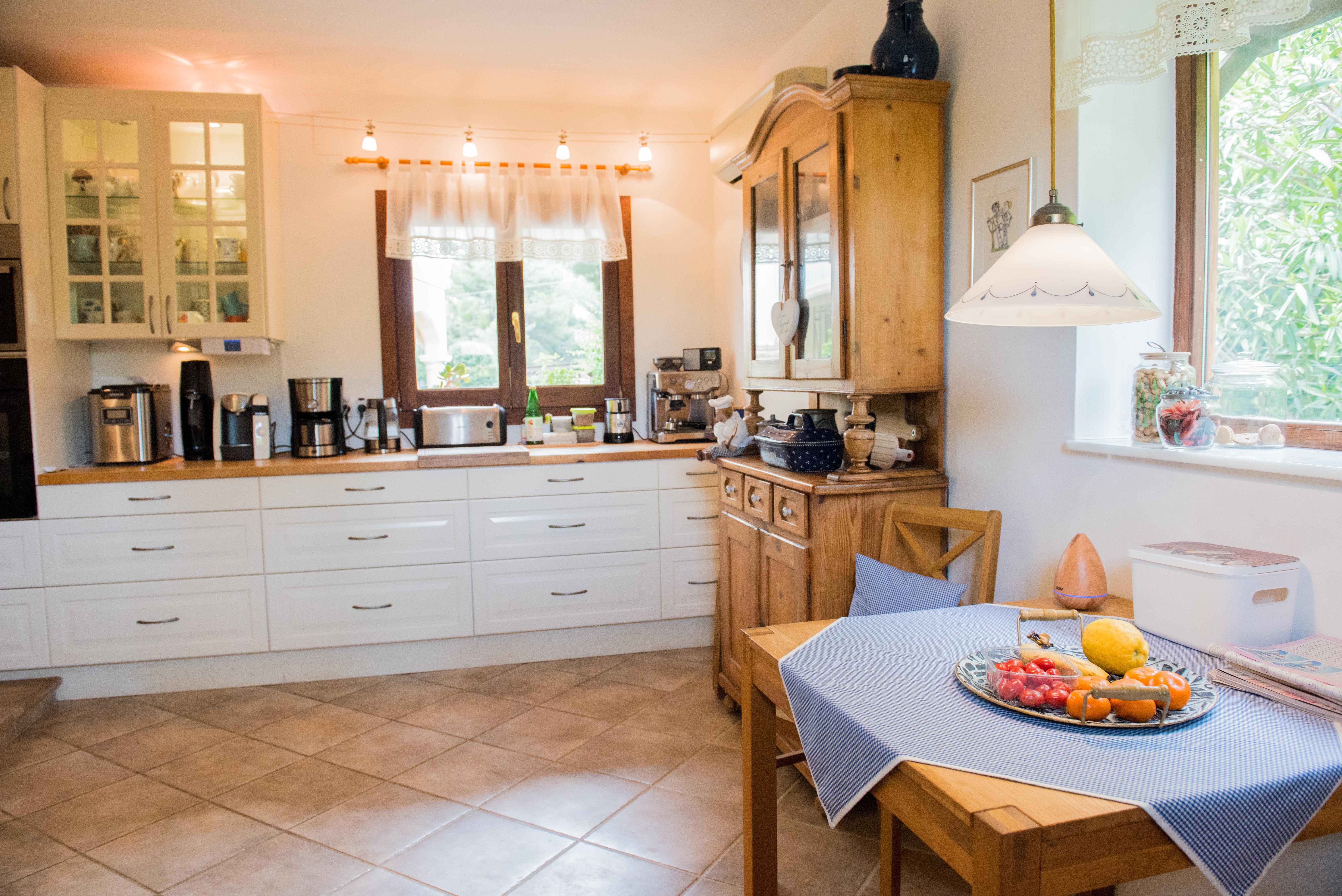 Rustieke keuken met witte kasten, houten werkbladen en eethoek. Inclusief een klassieke houten buffetkast, keramische tegelvloer en natuurlijk licht via twee ramen.