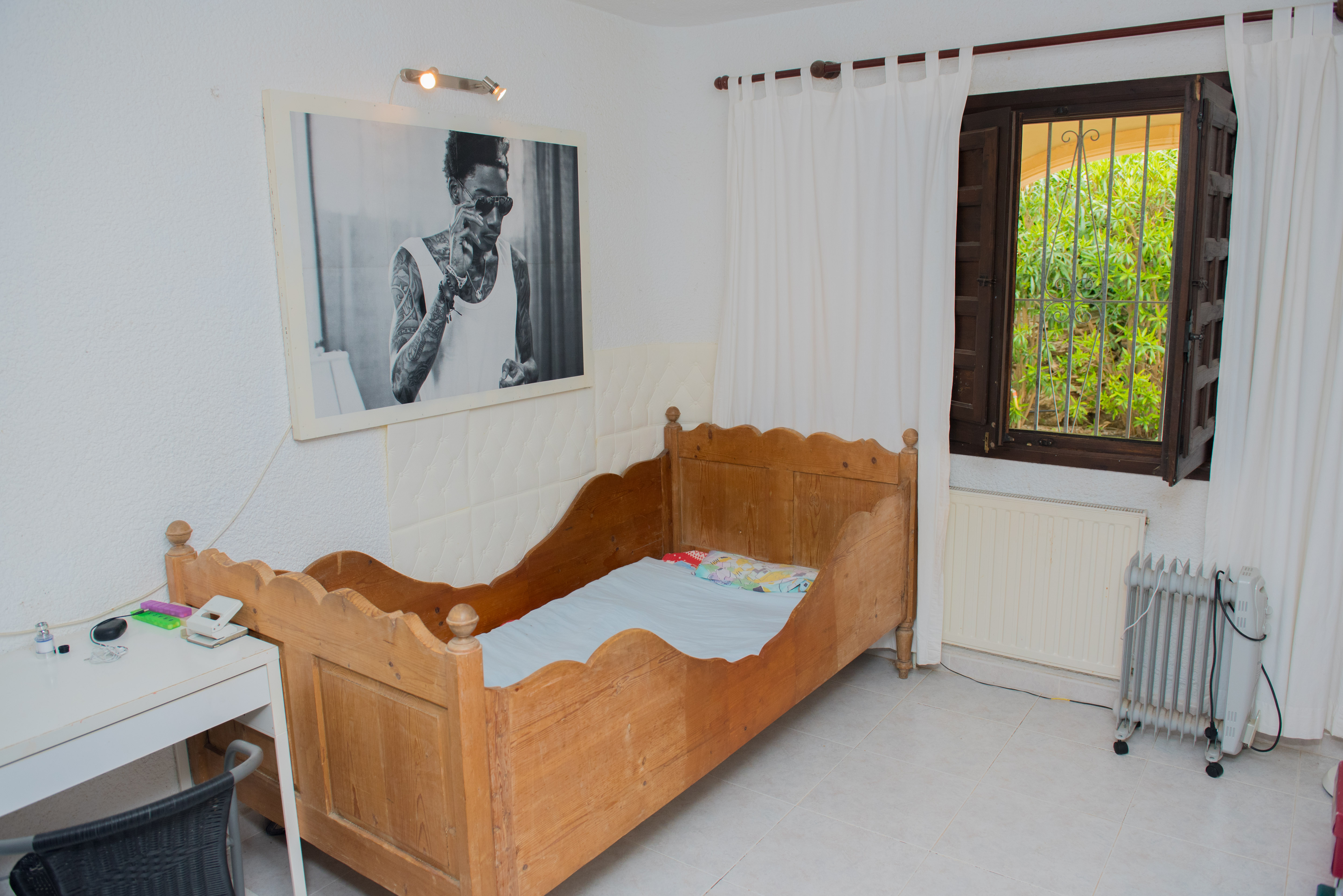 Slaapkamer met rustiek houten bedframe, wit bureau en donkerhouten raam met tralies. Voorzien van een lichte tegelvloer en uitzicht op de tuin.