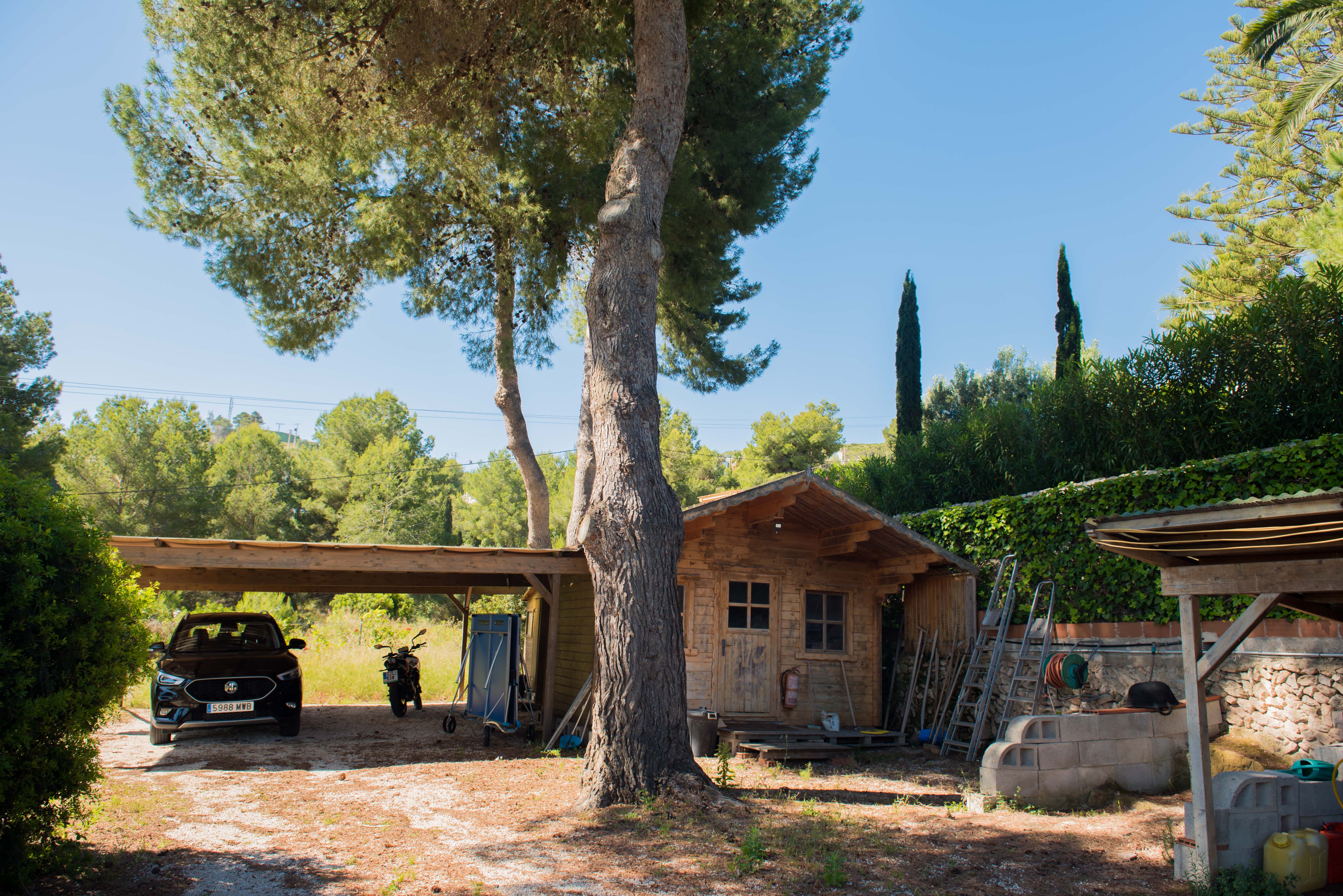 Uitzicht op de houten schuur en de overdekte garage, omgeven door mediterrane vegetatie en een heldere hemel.