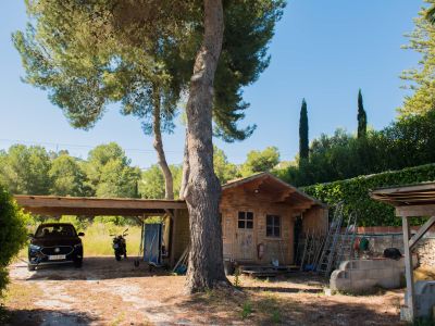 Uitzicht op de houten schuur en de overdekte garage, omgeven door mediterrane vegetatie en een heldere hemel.